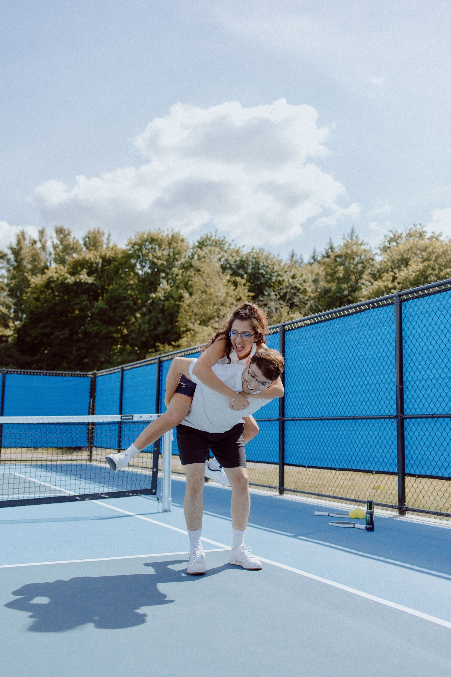 A playful moment captured on a bright blue tennis court surrounded by blue fencing and green trees on a sunny day.