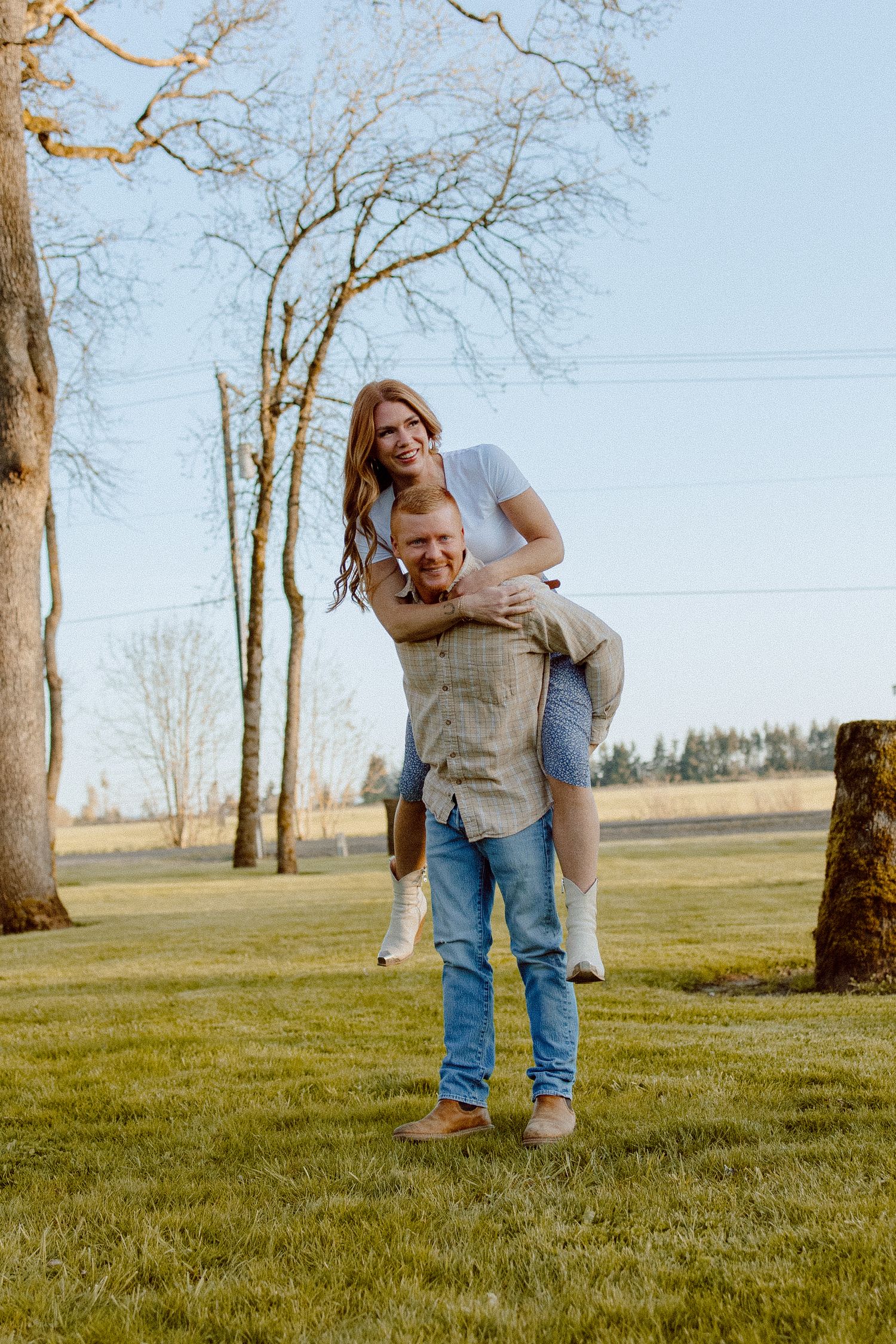 A playful couple sharing a piggyback ride moment in a grassy field with bare trees on a sunny day.