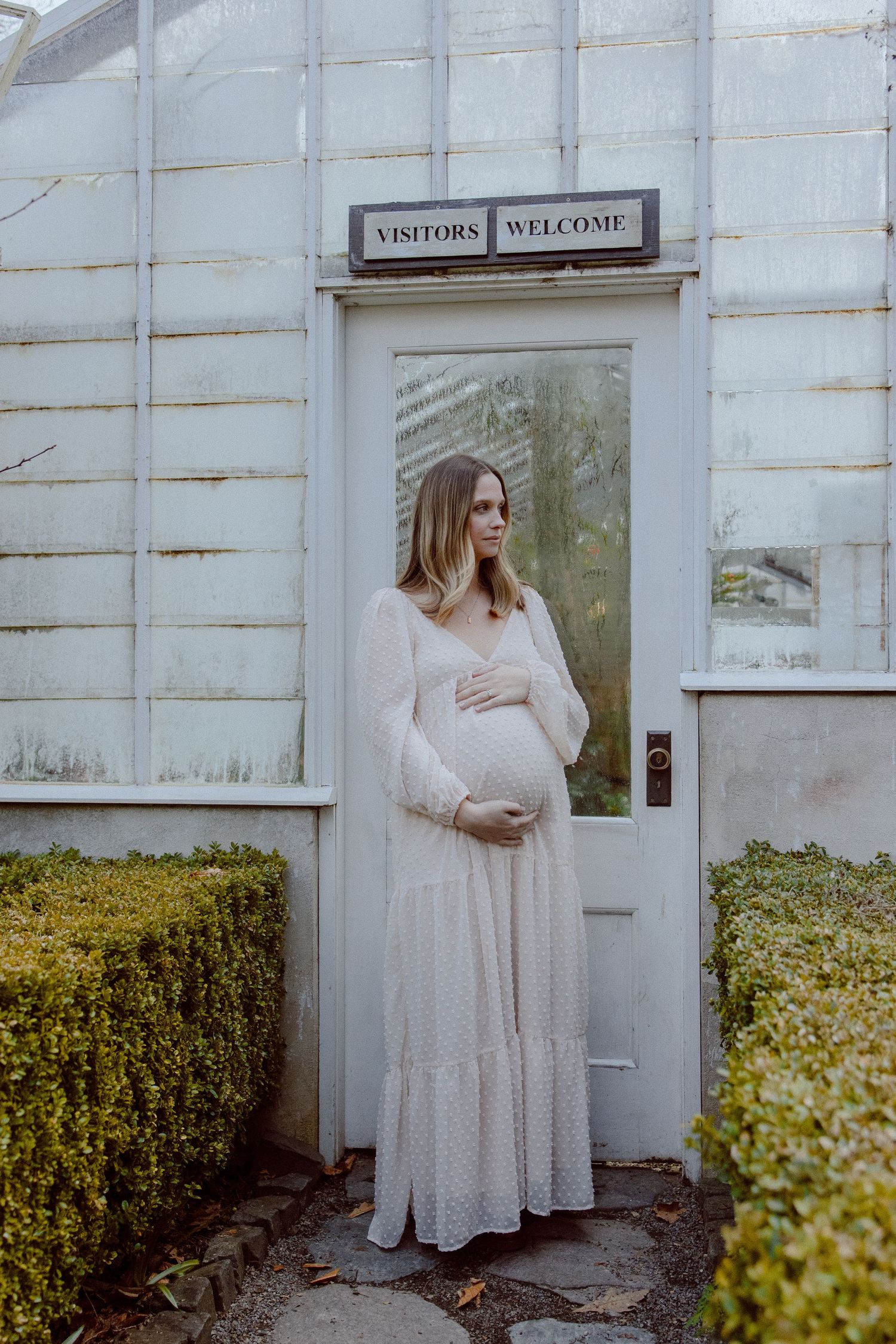 A pregnant woman in a flowing white dress stands in front of a greenhouse with a white door with a 'Visitors Welcome' sign above it.