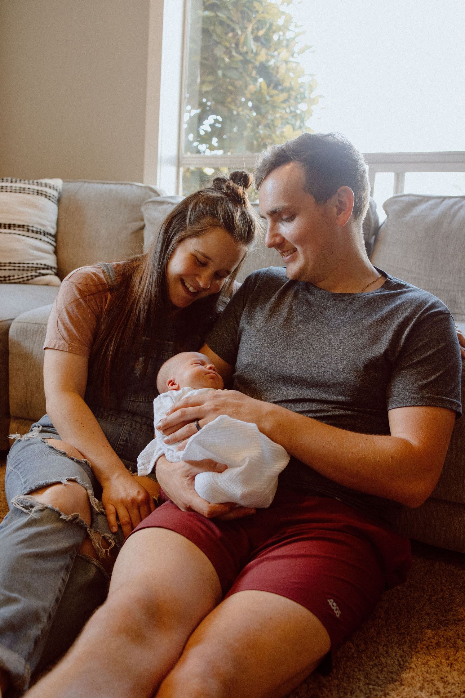Parents lovingly cuddle with their baby in front of a cozy brown couch in a sunlit living room.