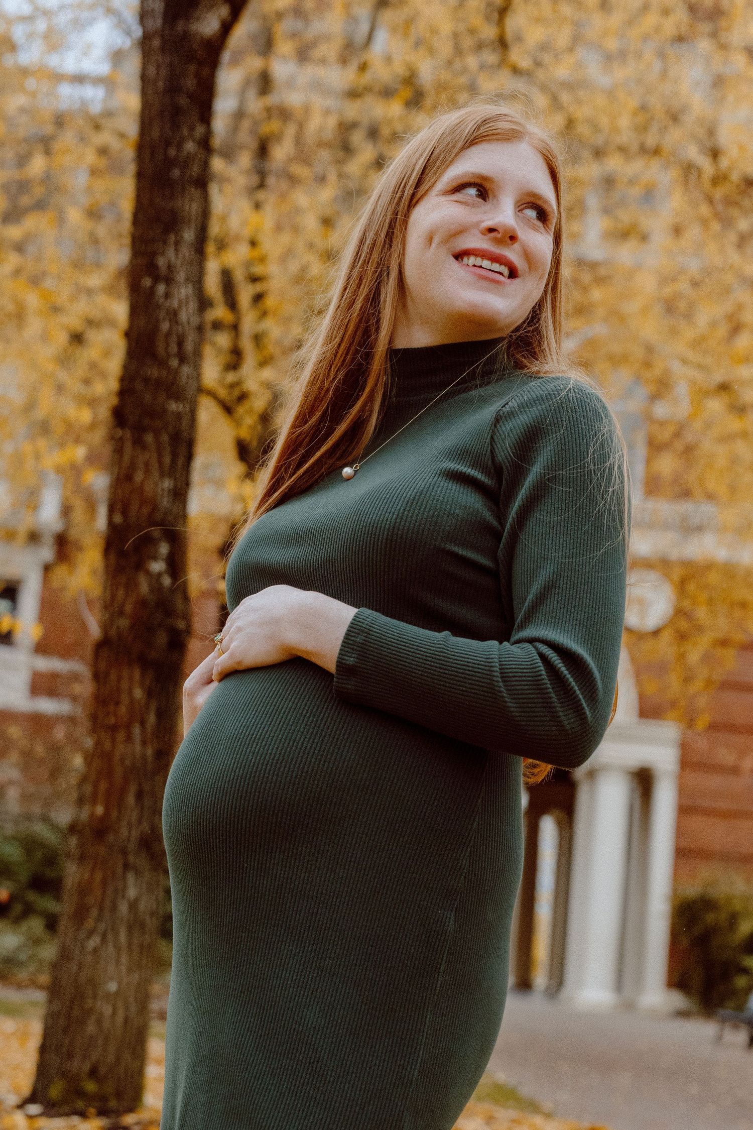 An expecting mother in a green sweater dress stands against fall foliage backdrop in warm autumn lighting.