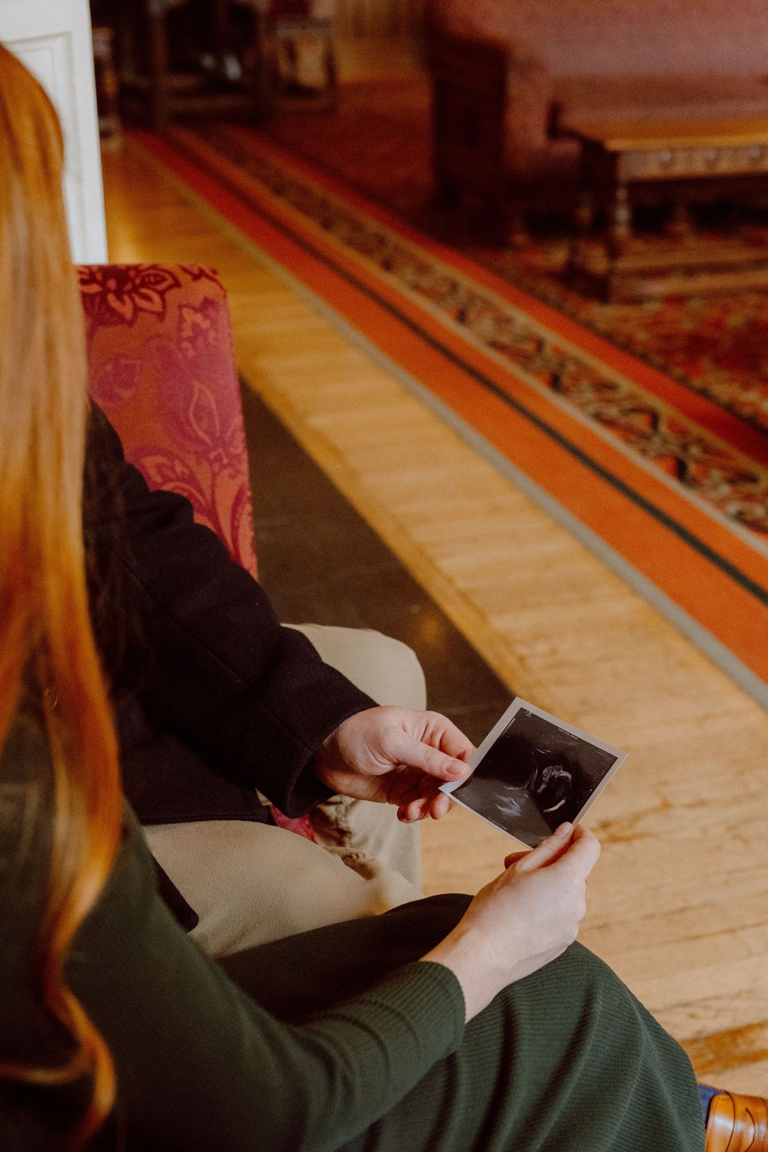 Hands in black sleeves hold a sonogram while sitting on a couch beside a patterned orange rug.