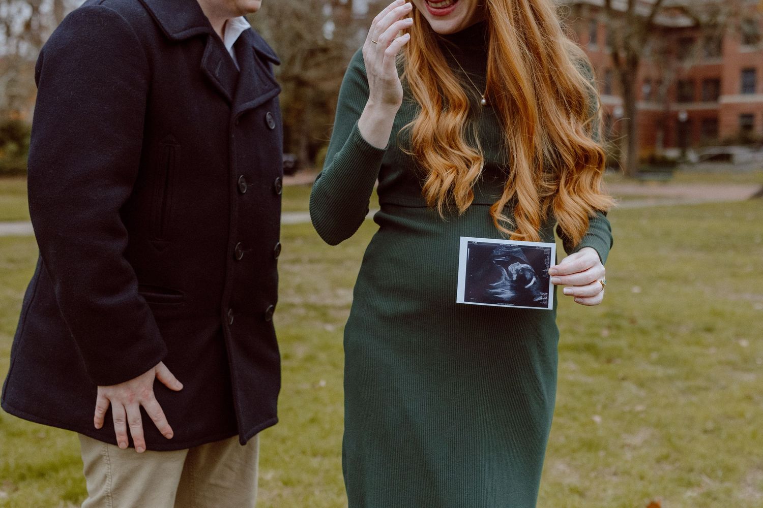 Professional outdoor photos showing a couple interacting playfully in a grassy area with their baby's sonogram photo.