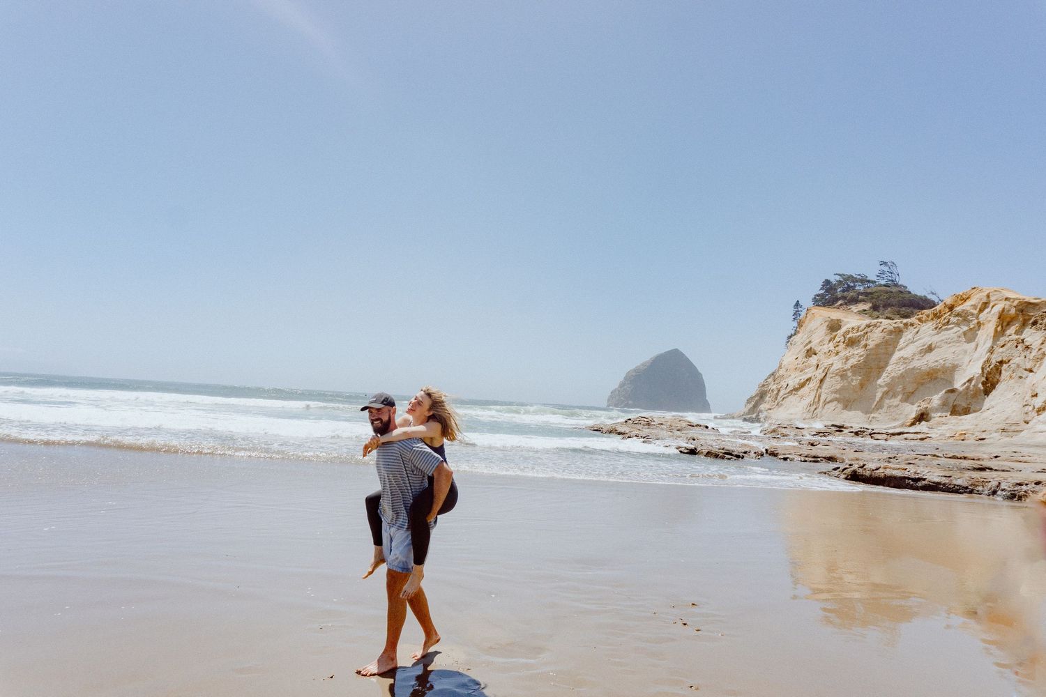 A piggyback ride on a sandy beach with dramatic coastal cliffs and rock formations in the background.