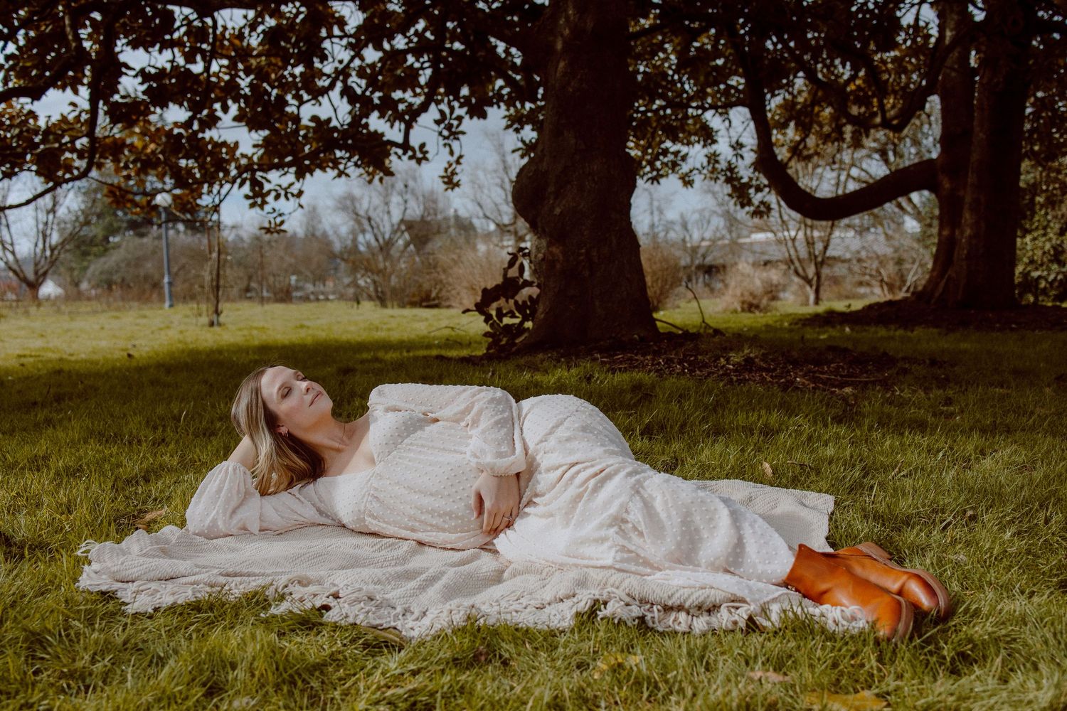 A pregnant woman in a flowing white dress lays down in a grassy field during a dreamy outdoor photo session.