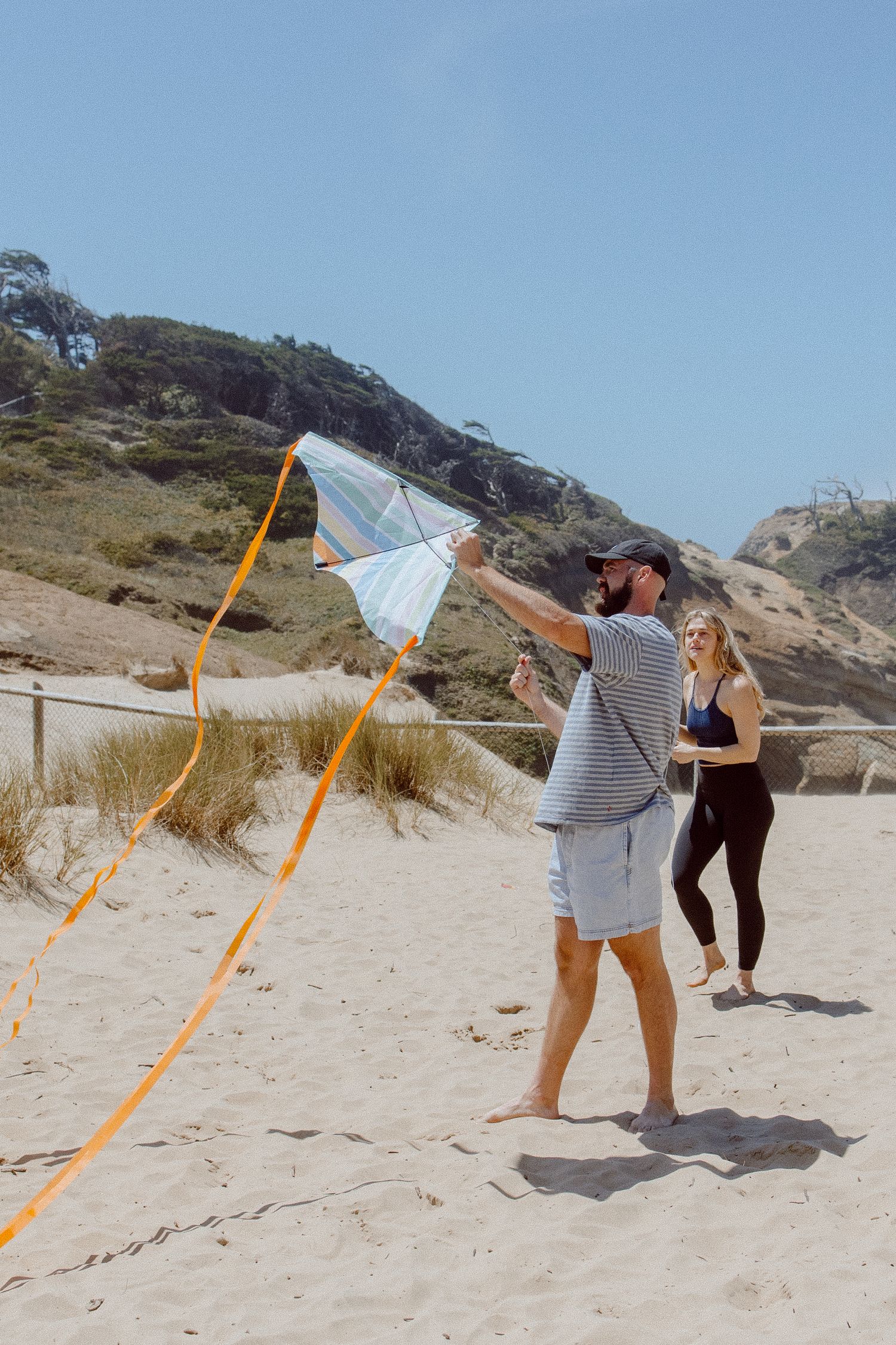 A sequence of beach scenes showing a winged kite structure being assembled in five progressive stages on sandy terrain.