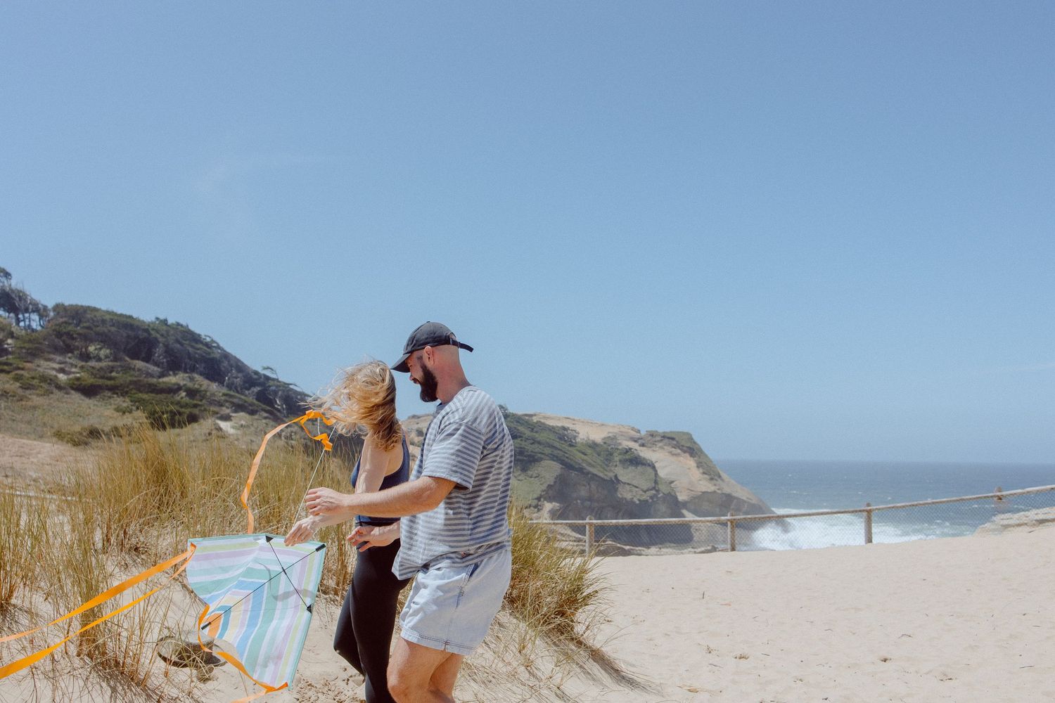 Couple stands with a kite on scenic coastal sand dunes overlooking a beach on a sunny day.