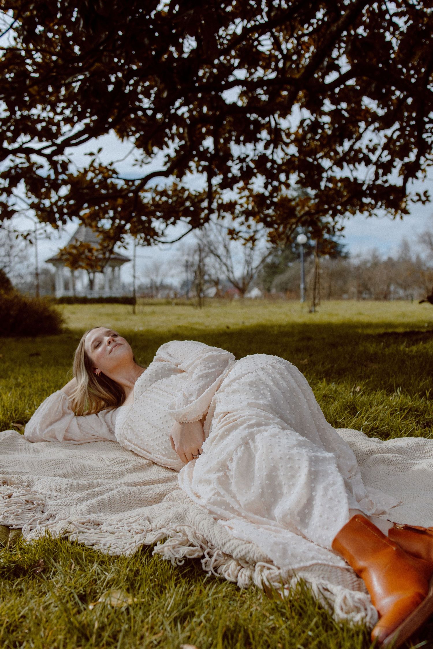 A person in a flowing white lace dress lies peacefully on a blanket in a grassy field beneath blooming trees.