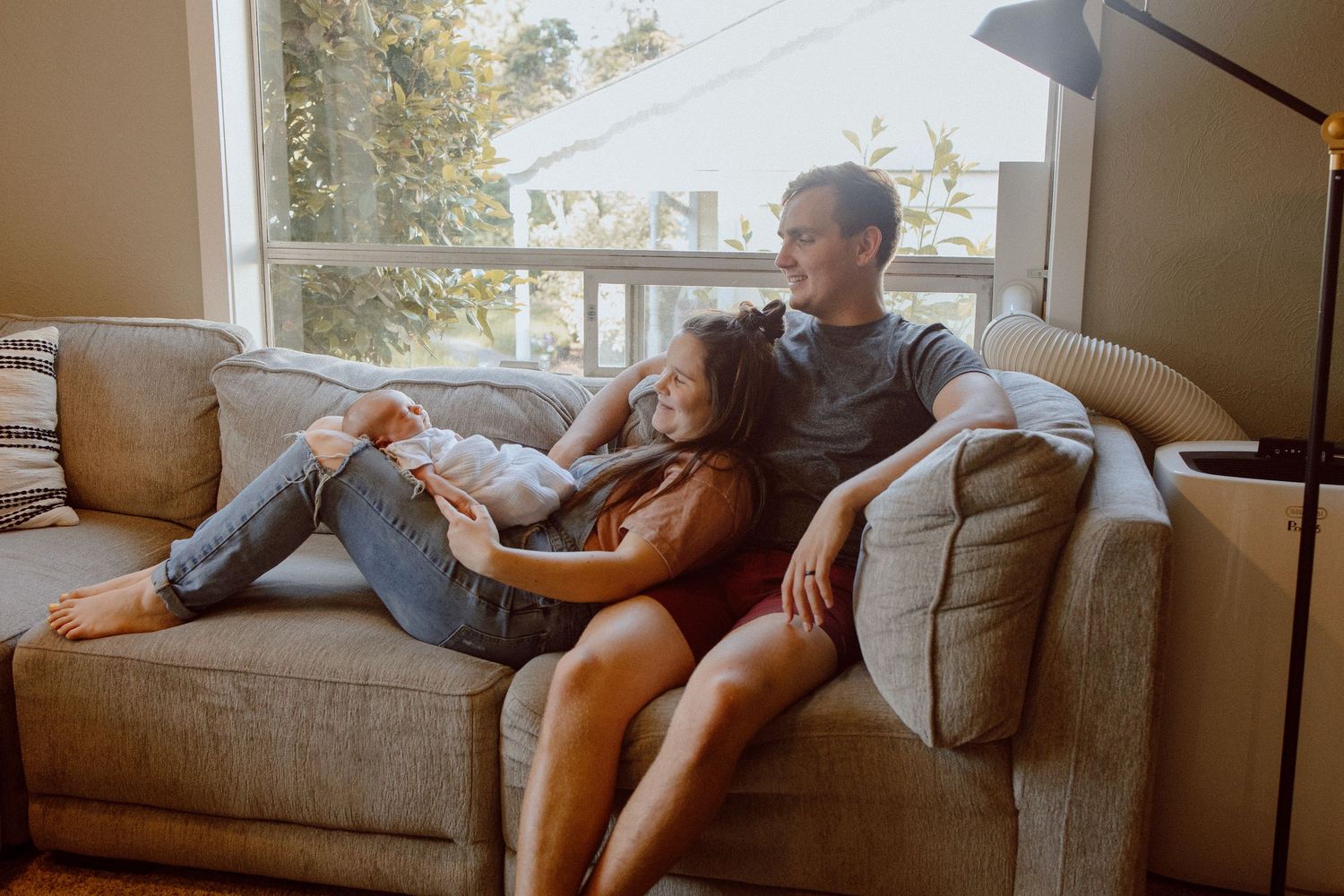 A sequence of photos showing a couple lounging and relaxing with their newborn baby on a brown couch in front of a big window.