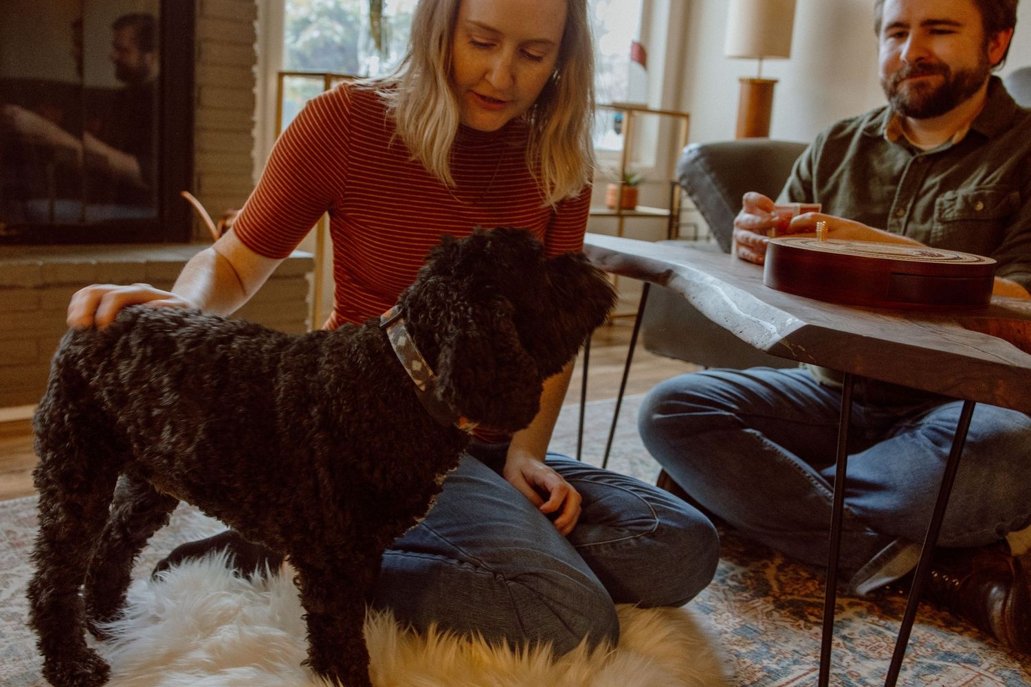 A couple gathered in the living room with their black dog giving it snuggles while they play a board game.
