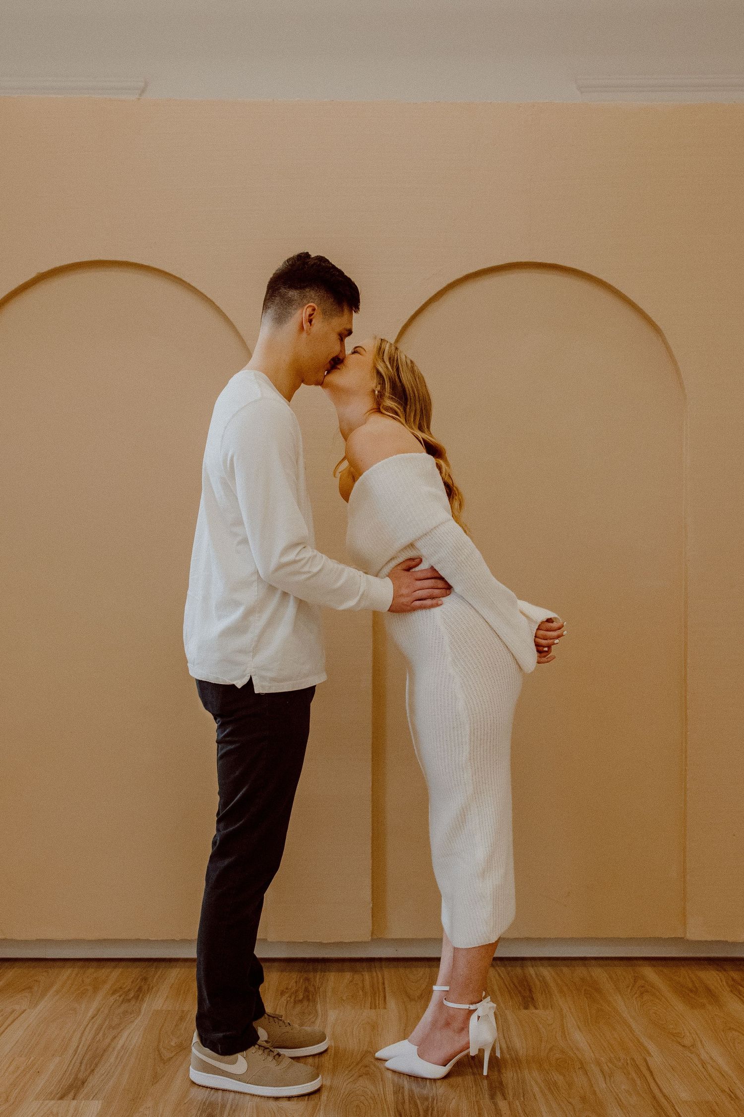 Two people in white and dark attire embracing in a minimalist room with wooden floors and neutral arched walls.