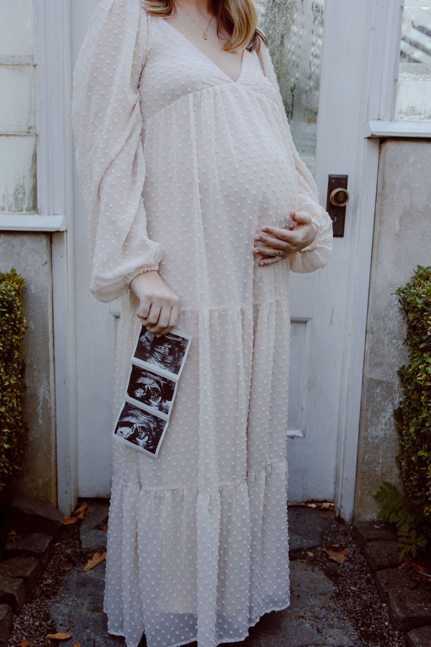 Pregnant woman in white flowy maxi dress holds ultrasound photos while standing by rustic door outdoors.