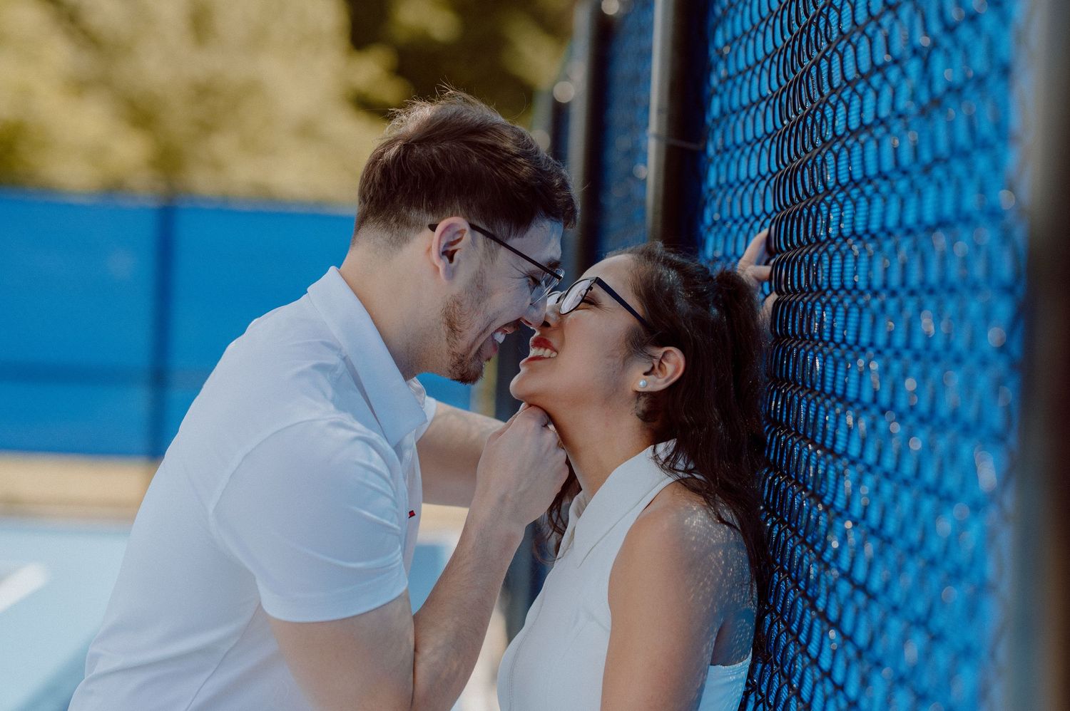 Romantic couple sharing an intimate moment against a bright blue wire fence background of a tennis court on a sunny day.