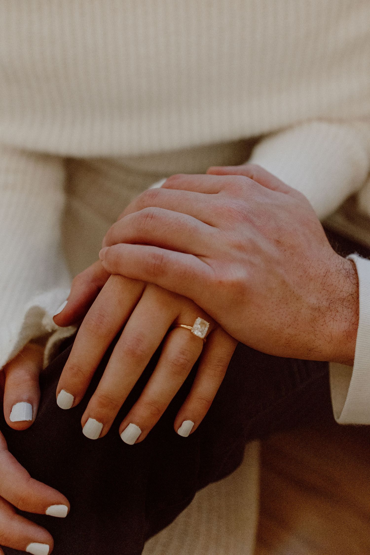 Two hands tenderly touch with one wearing white nail polish and a square-cut diamond engagement ring.