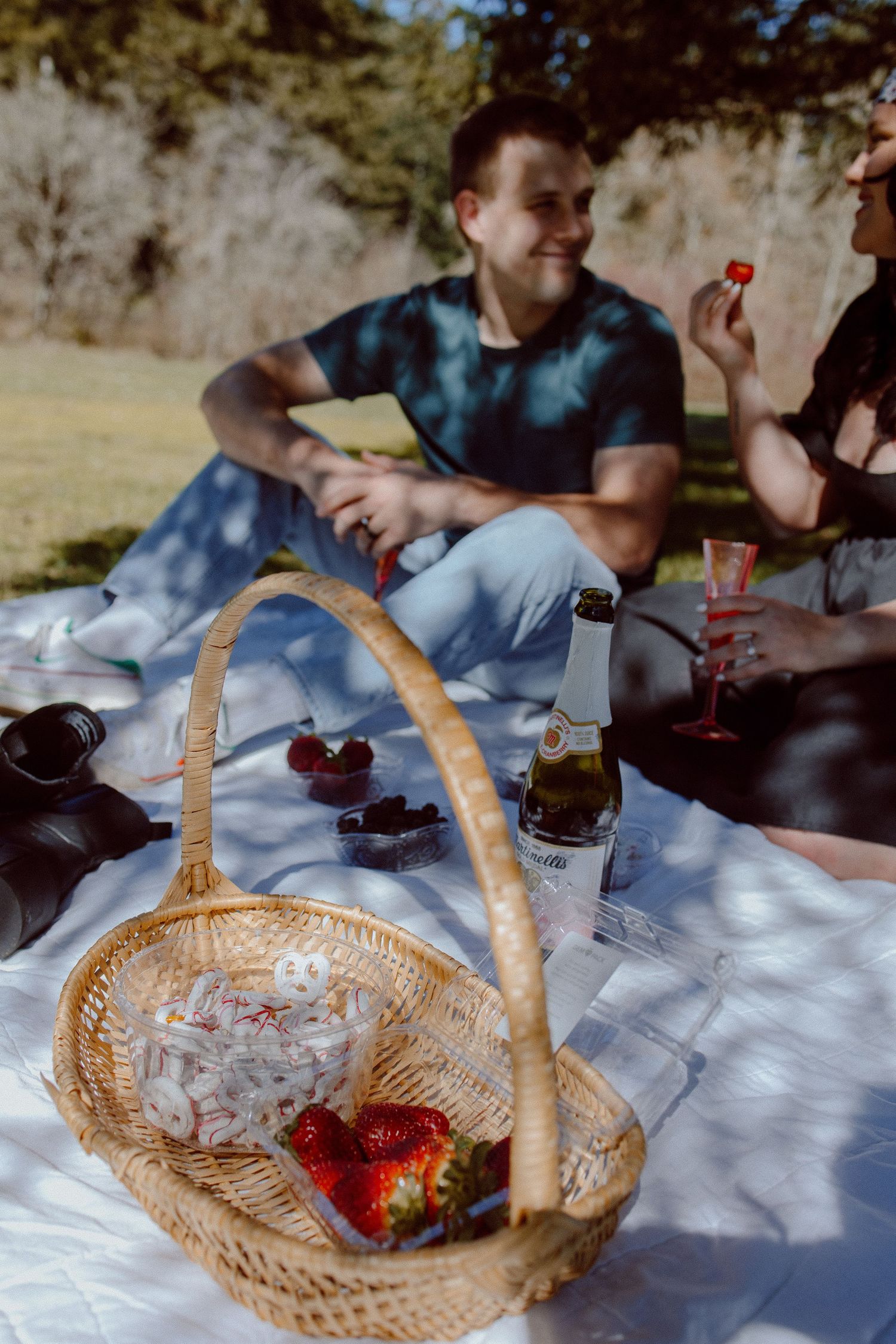 A wicker basket with strawberries sits on a picnic blanket as a couple enjoy drinks and snacks outdoors on a sunny day.