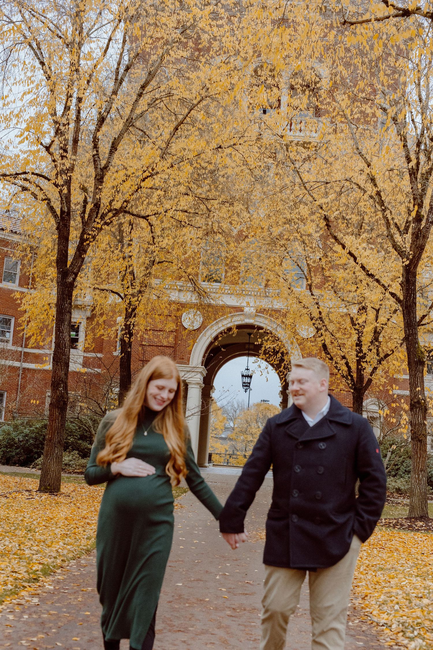 A couple walks hand in hand beneath golden autumn trees on a brick pathway with an arched entryway behind them.
