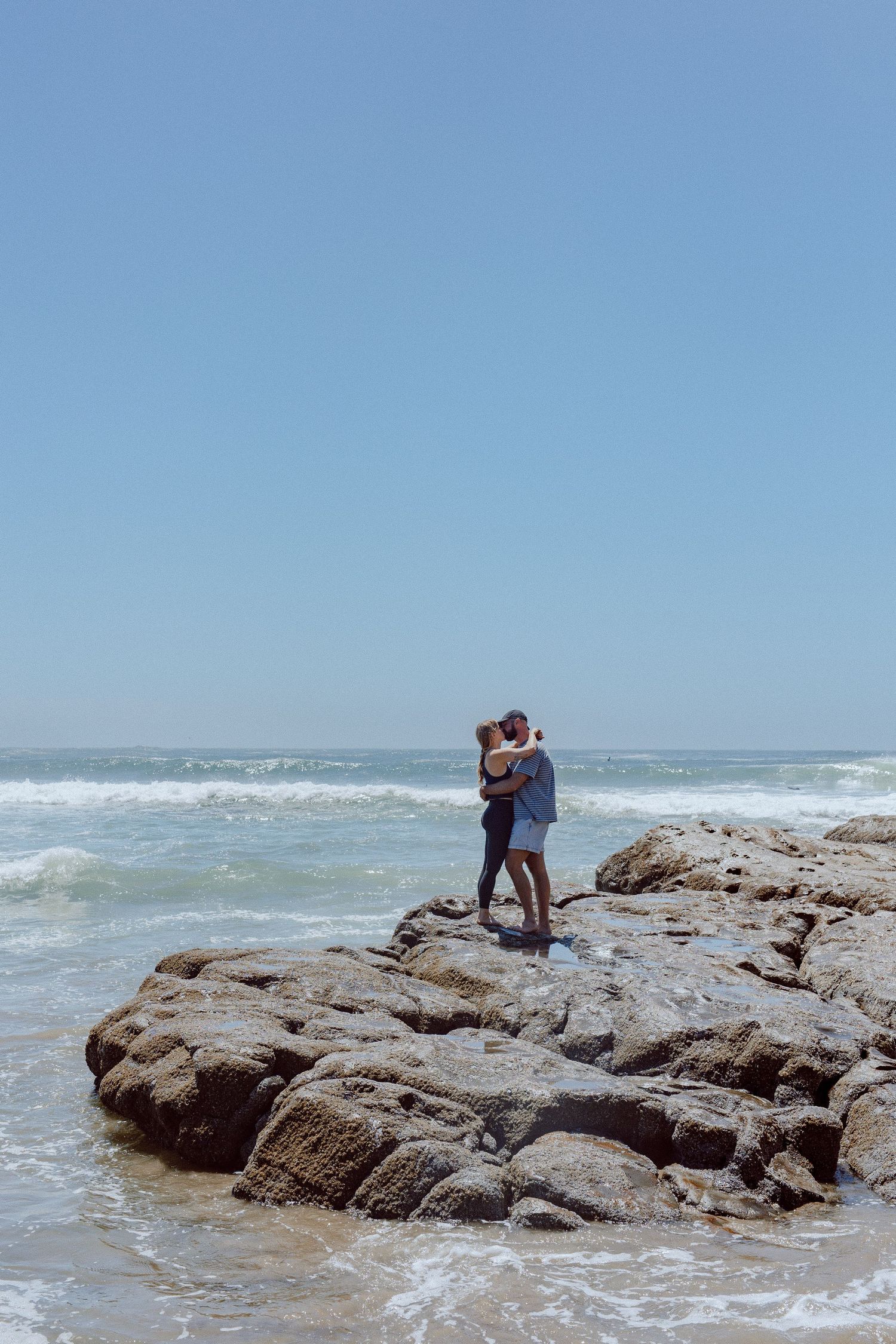A couple stands intimately on rocky coastal outcrop as ocean waves crash against the shore on a bright sunny day.