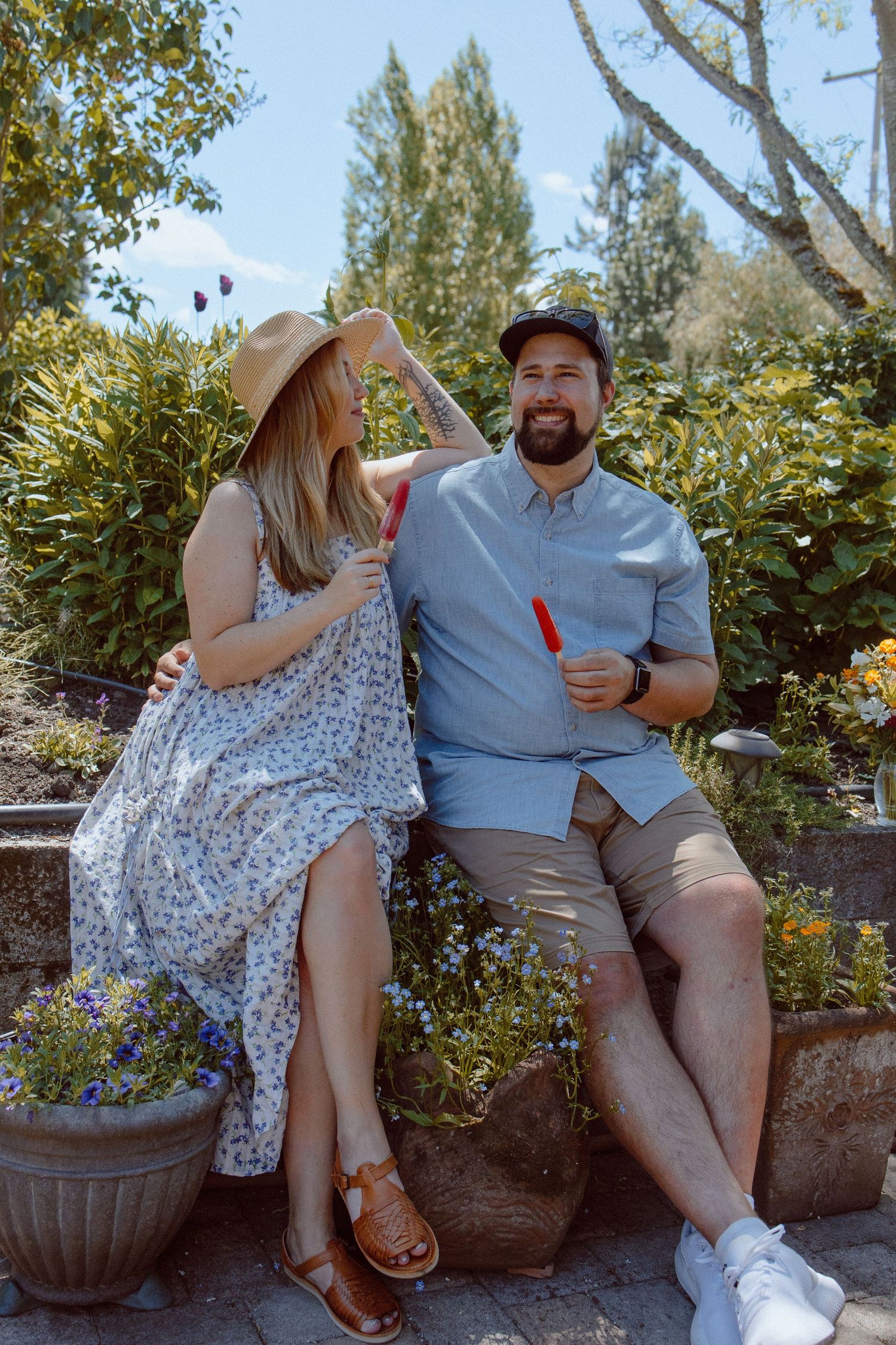Couple sitting casually together on stone wall in sunny garden with large decorative planters sharing a popsicle stick.