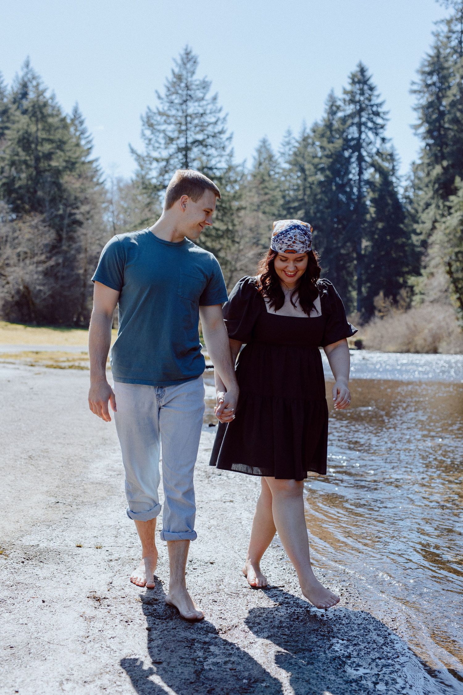 Two people stroll along a river beach on a sunny day with tall evergreen trees in the background.