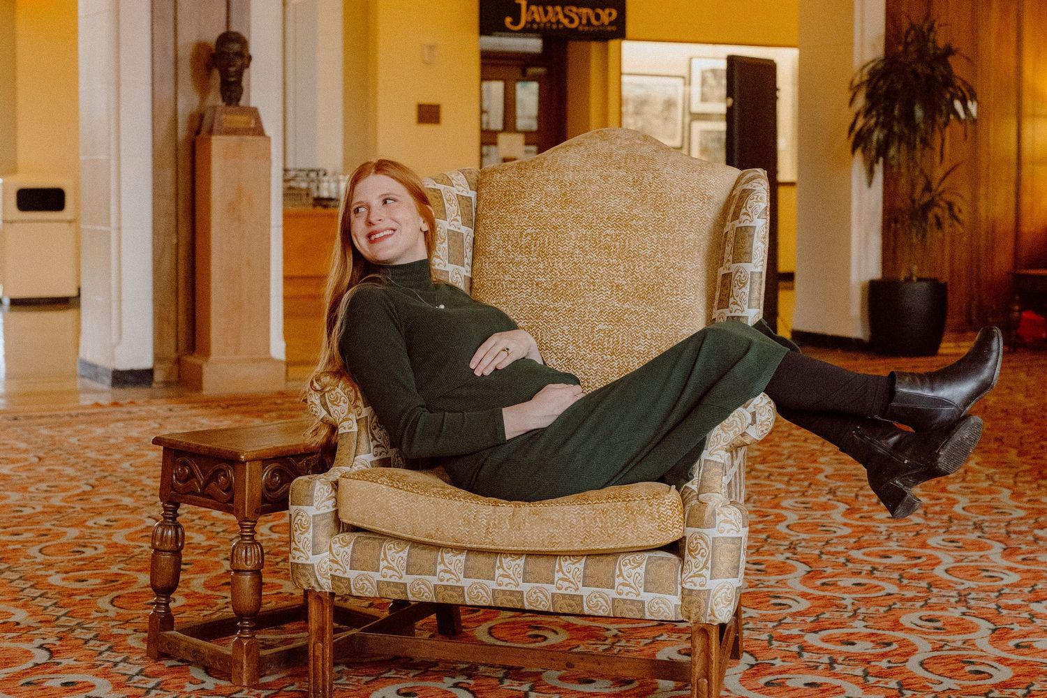 A candid lounging shot of a mother in an antique chair against ornate orange and brown patterned carpeting in a vintage-style interior.