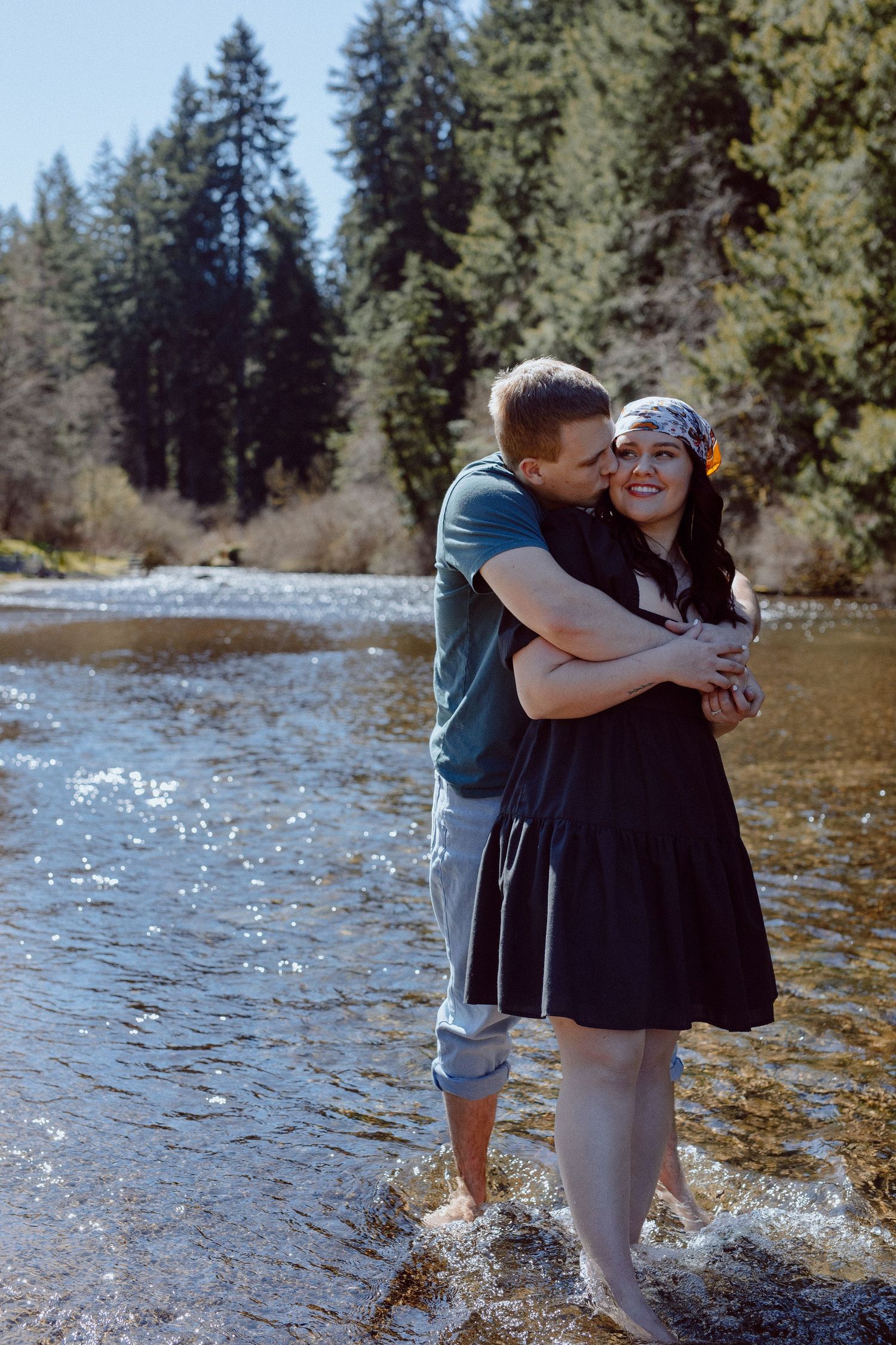 A couple embraces and shares a sweet moment while they stand in a scenic river surrounded by evergreen trees.
