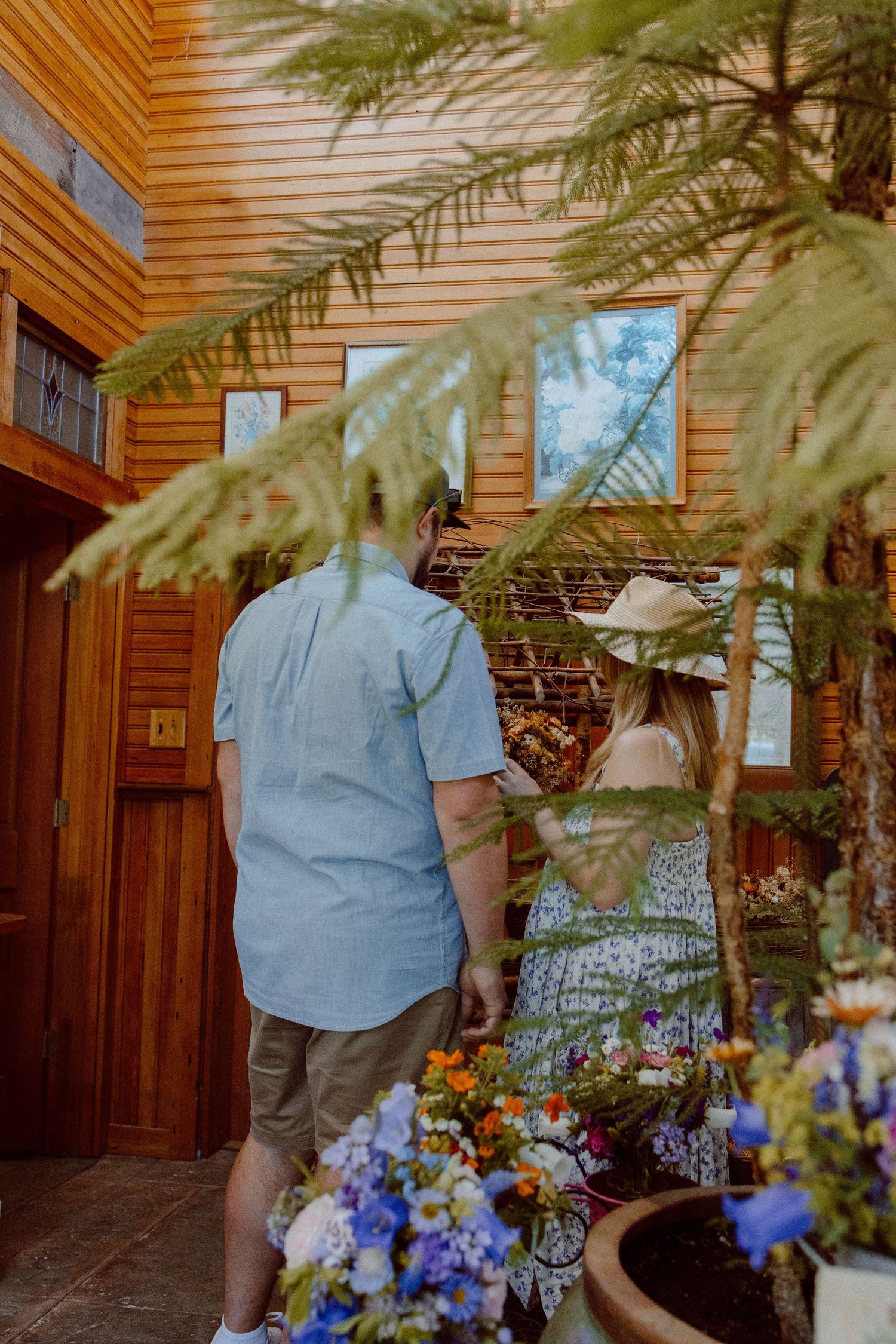 A giant potted tree sits in the middle of a flower shop next to colorful blue and yellow flowers in an indoor setting.