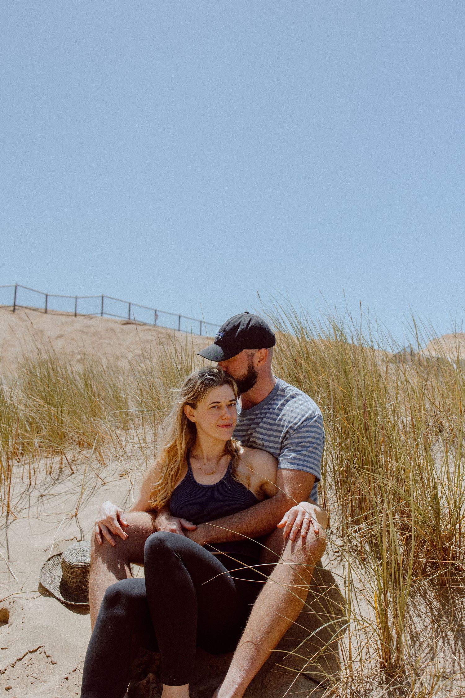 A couple sits together intimately on sand dunes with beach grass and blue sky in the background.