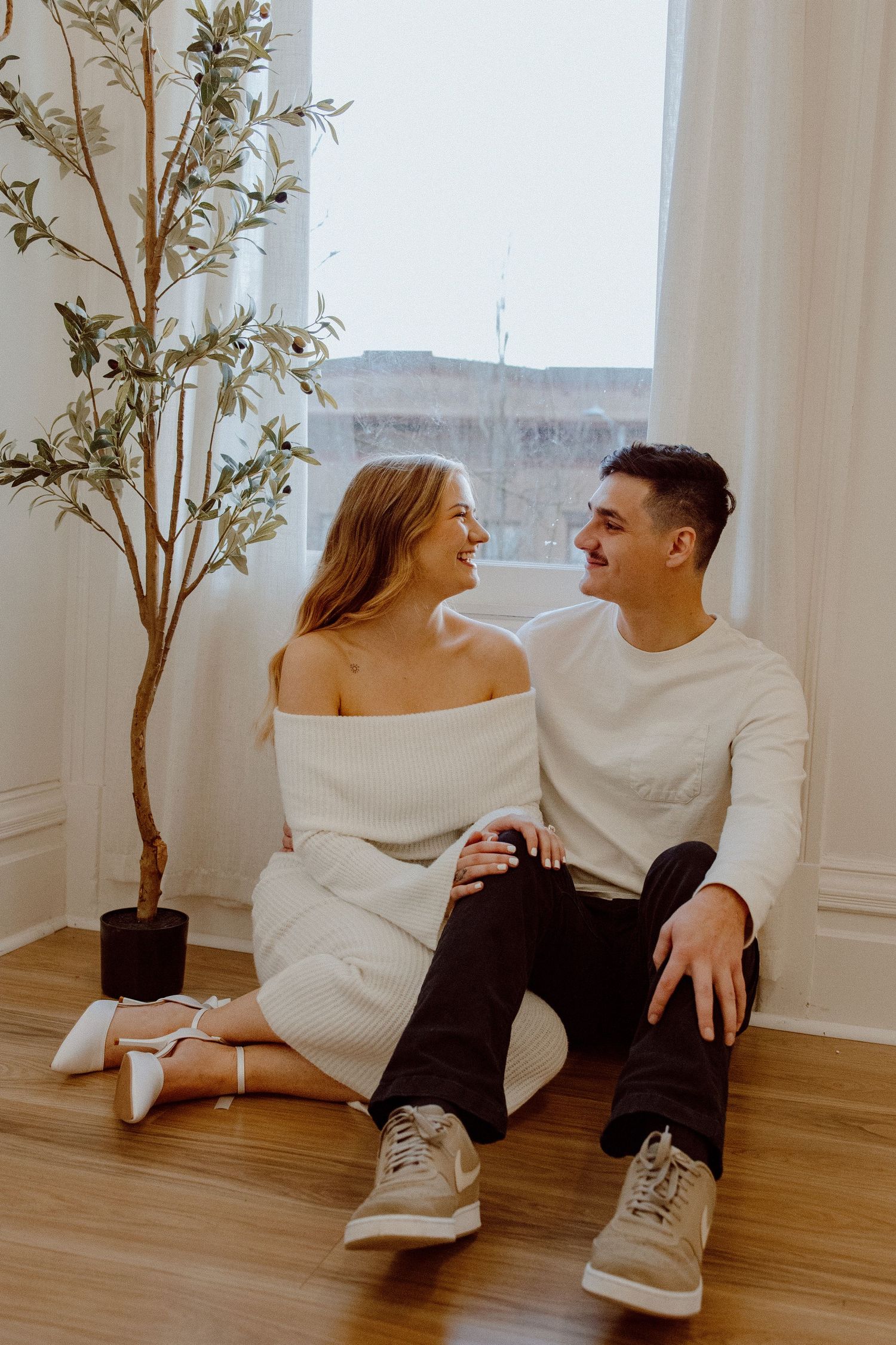 A romantic couple in stylish white clothing shares laughter while sitting together on a hardwood floor.