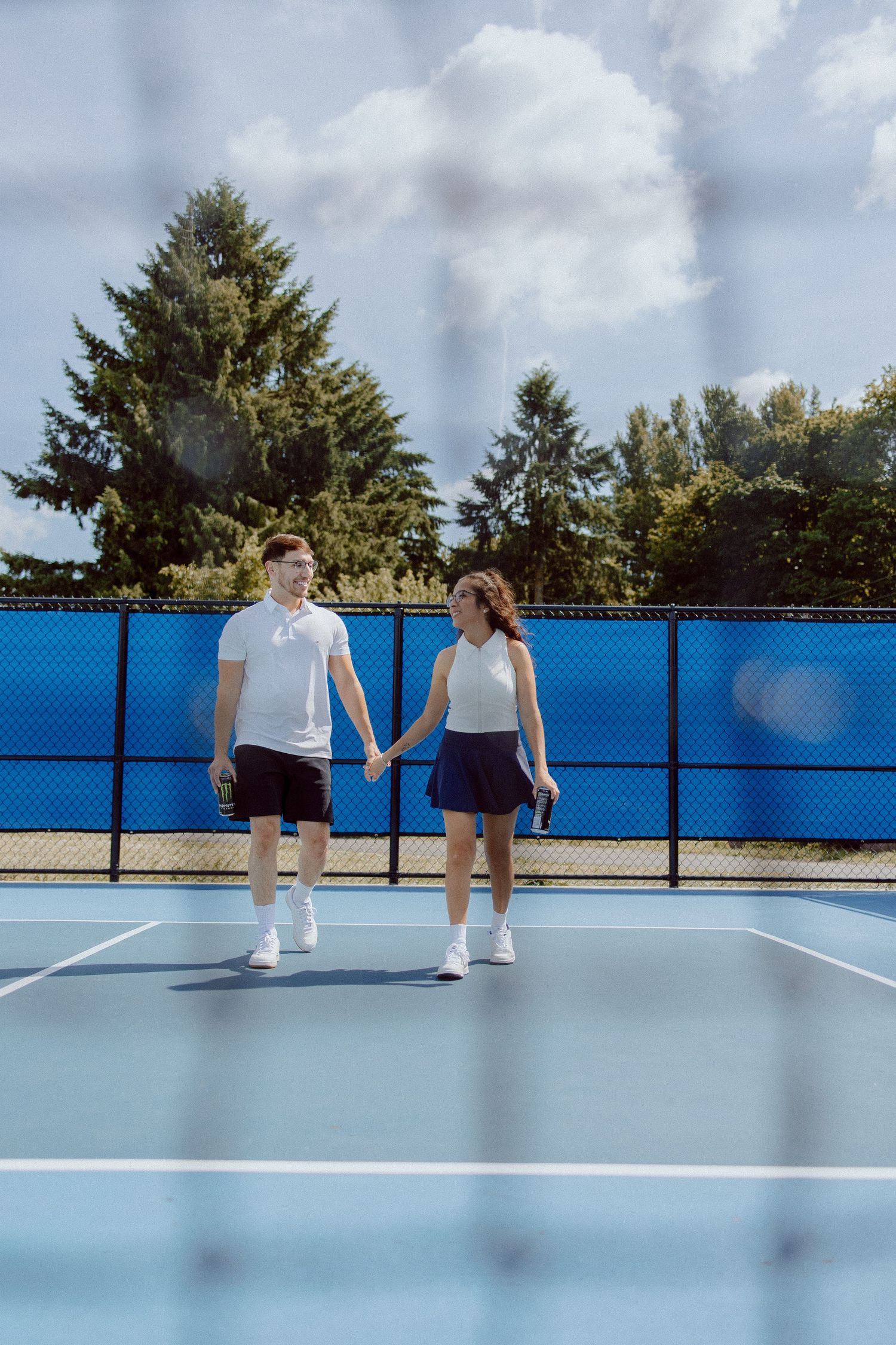 Two people standing on a bright blue tennis court under cloudy skies as trees line the background.