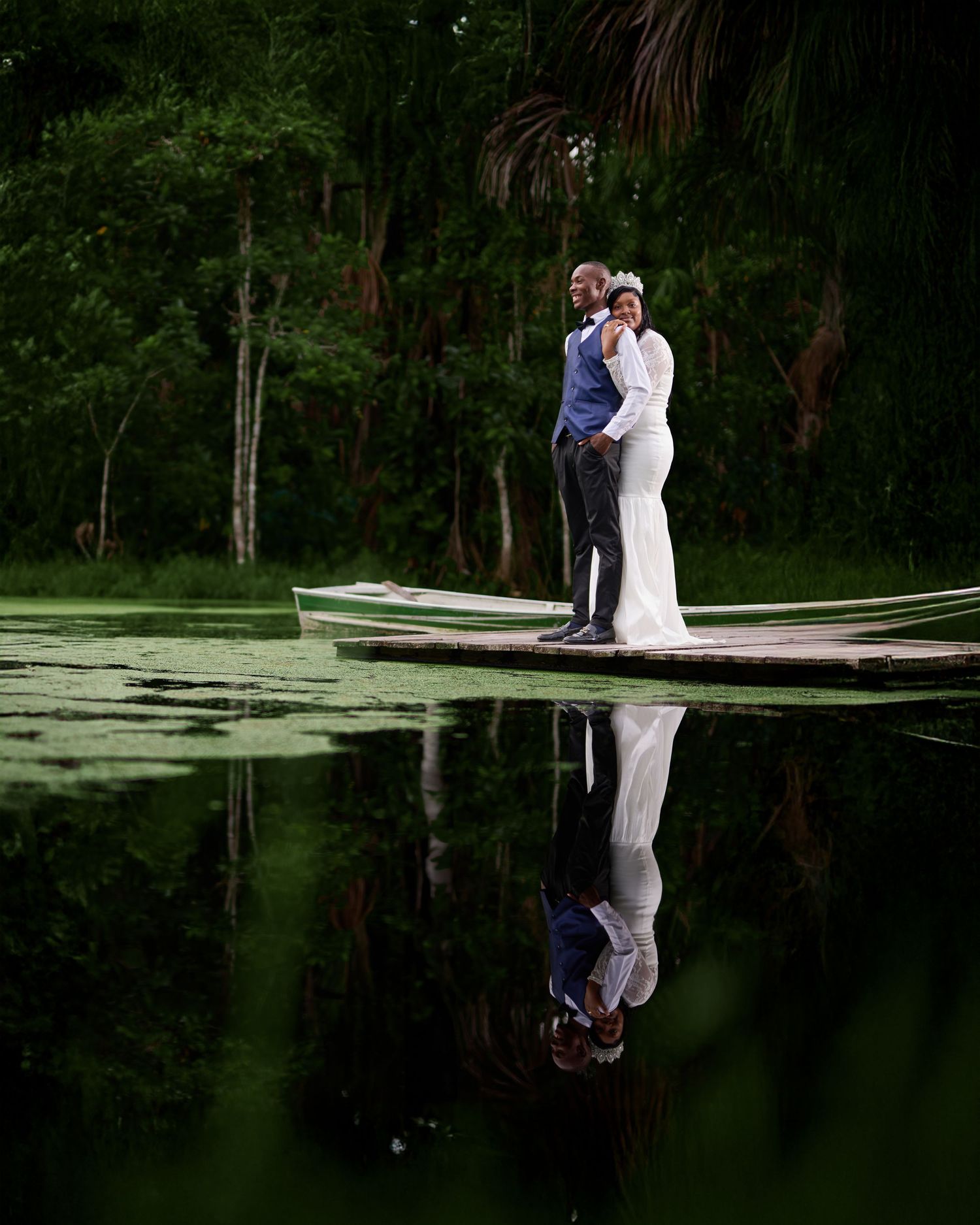A couple shares a romantic moment on a wooden dock overlooking a serene lake surrounded by trees at sunset.