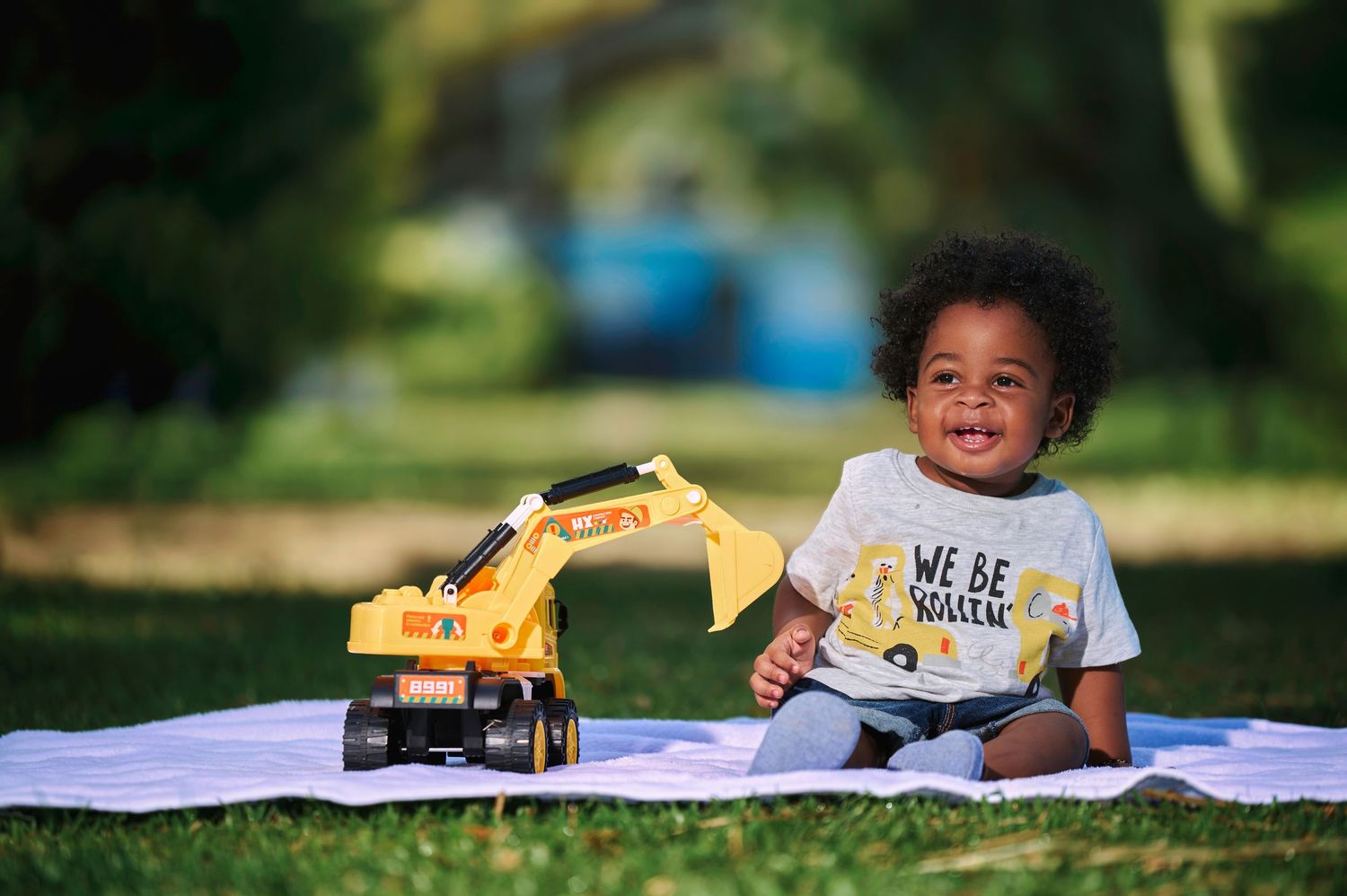 Child lying on white blanket outdoors playing with yellow toy trucks.