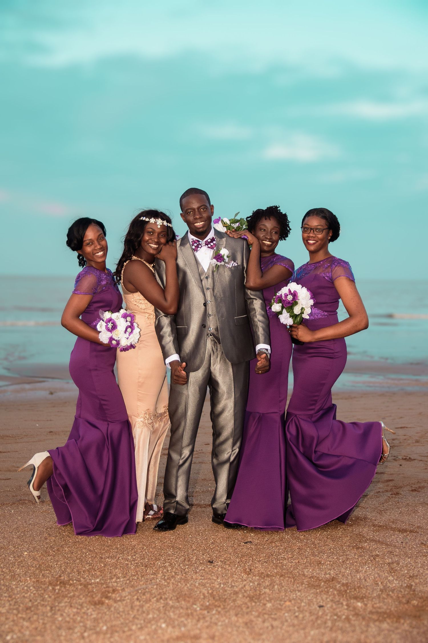 Wedding party in purple and gray attire poses together on a beach with turquoise sky.