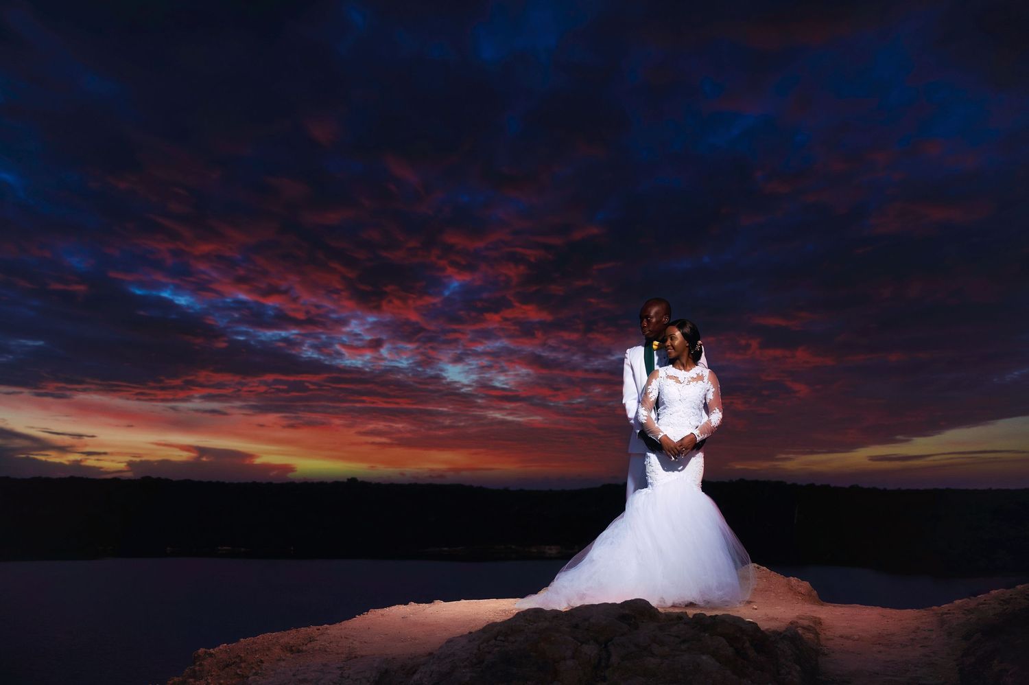 Wedding couple embraces on cliff edge under vibrant purple and red sunset sky.