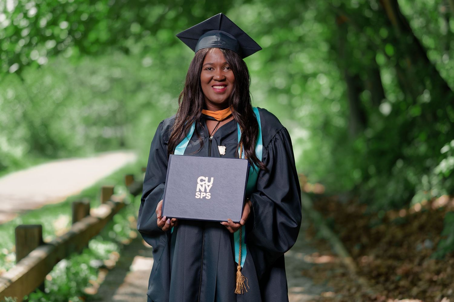 Graduate in cap and gown poses outdoors against lush green background.