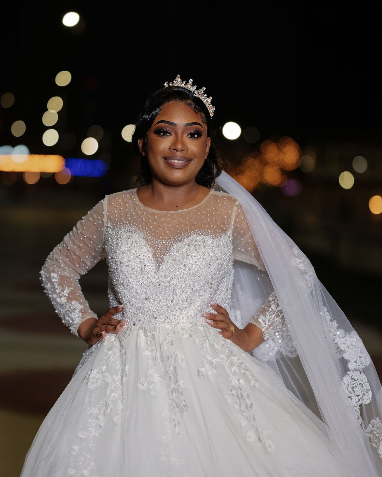 Elegant bride in a beaded long-sleeve ball gown with tiara and veil posing outdoors at night.