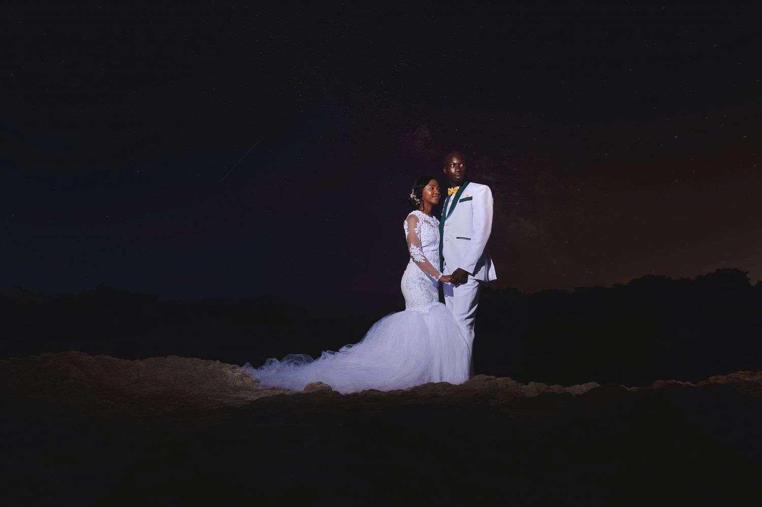 Couple in wedding attire poses romantically under a starry night sky outdoors.