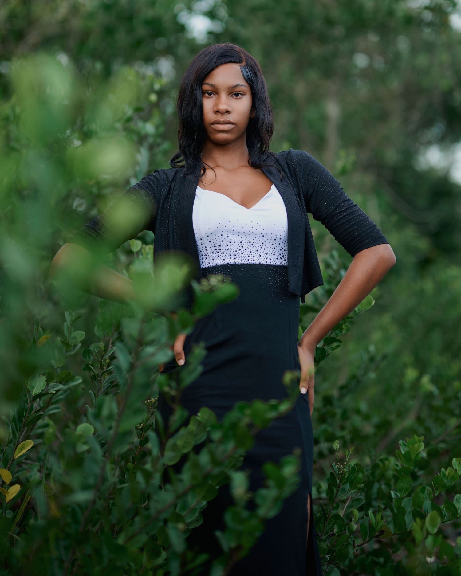 Person in black cardigan and white top poses among lush green foliage.