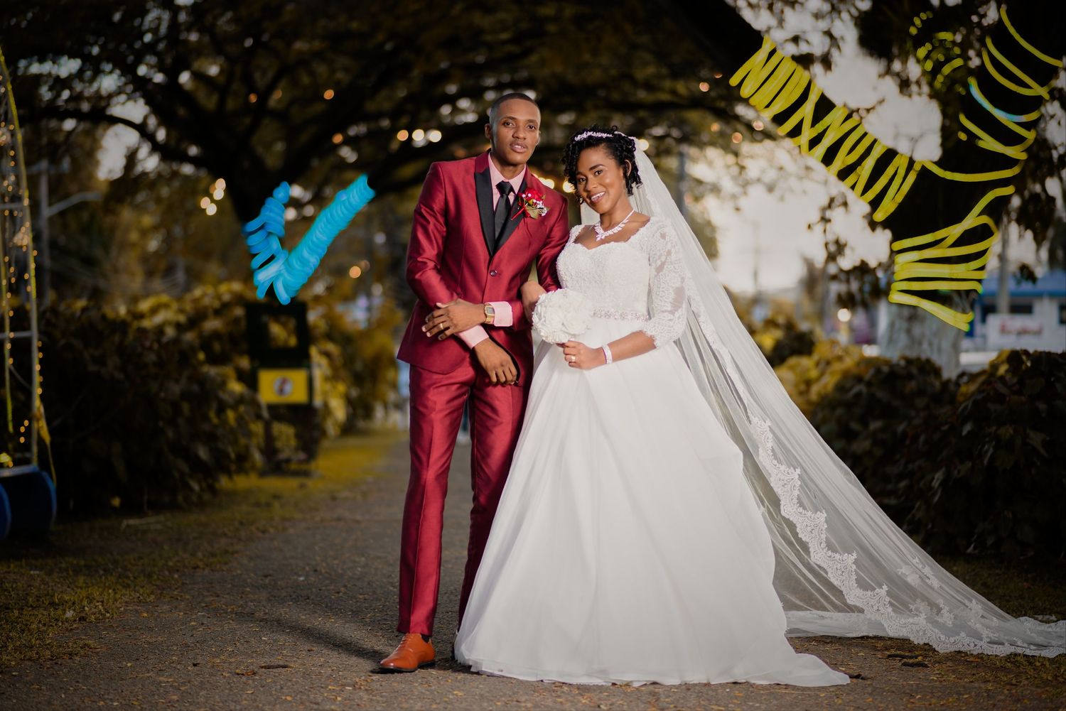 Elegant bride in white lace gown and groom in red suit posing together at an outdoor wedding celebration at night.