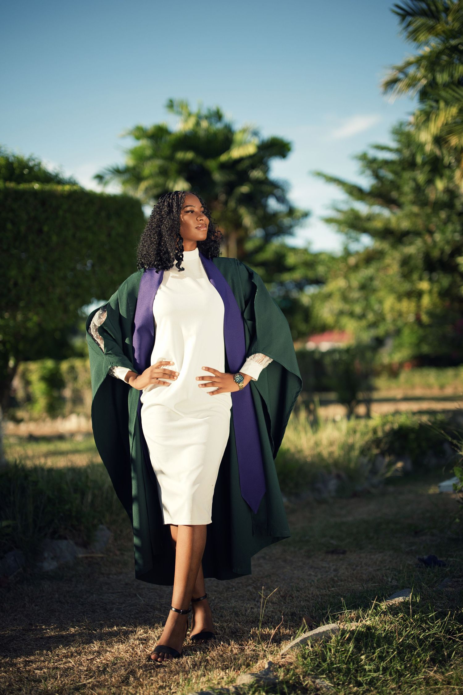 Graduate in academic regalia poses against natural outdoor backdrop with palm trees.
