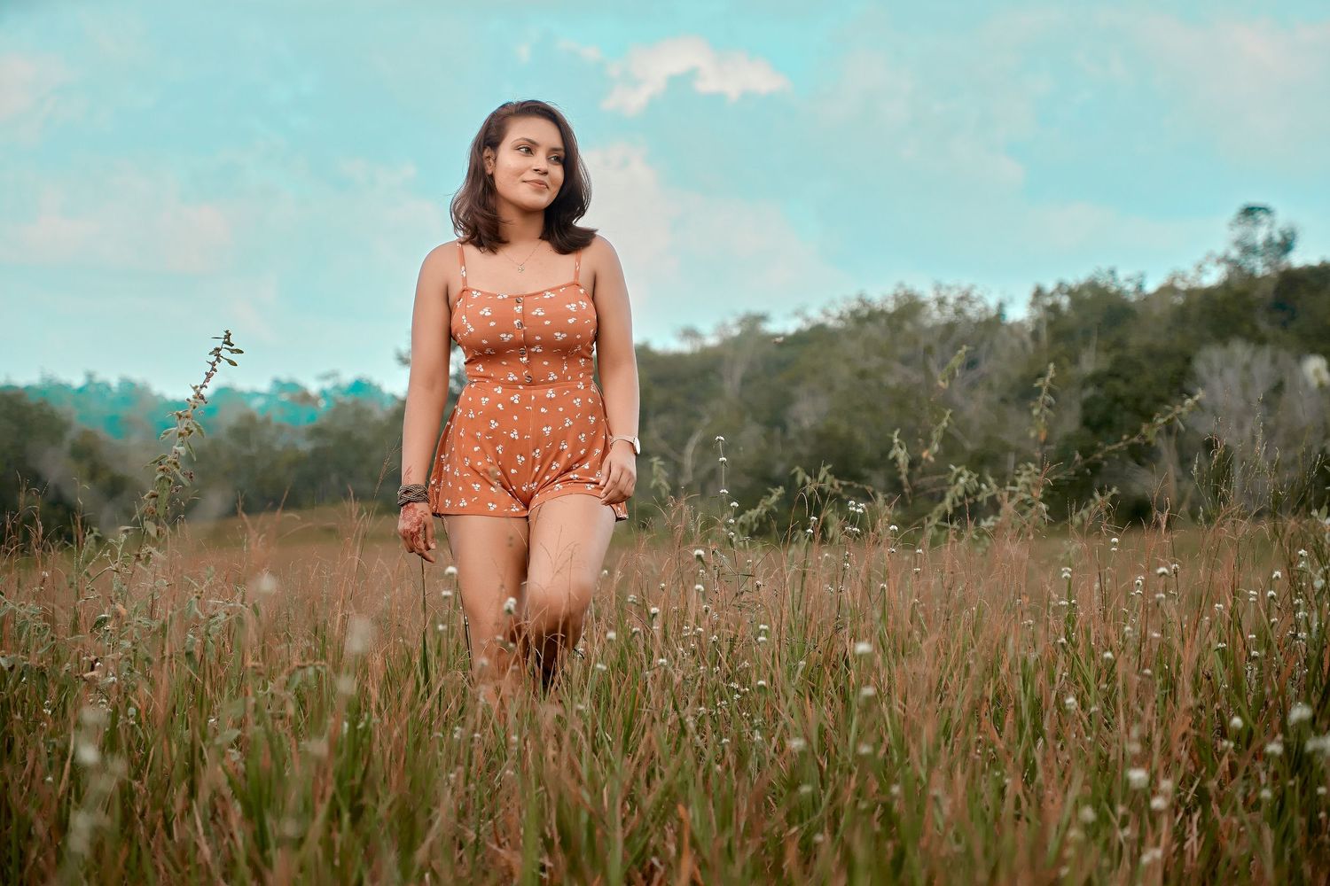 A person in an orange floral dress poses gracefully in a sunlit meadow with tall grass and blue skies.