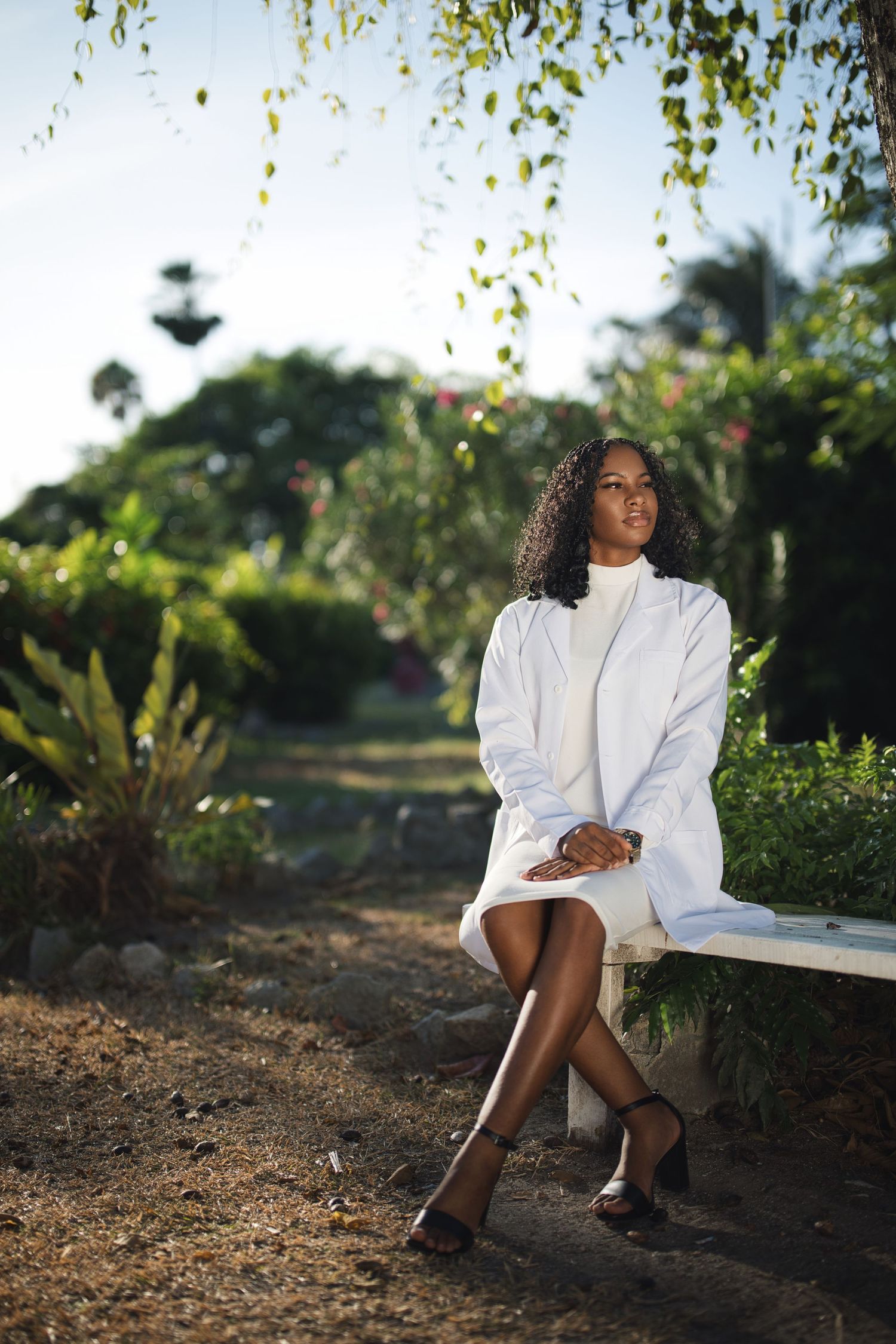 A young Black woman in a white dress and lab coat sits on a bench in a lush tropical garden.
