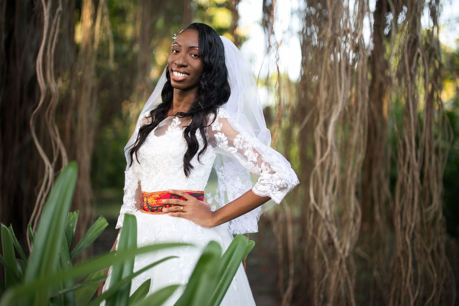 Smiling Black bride in white lace gown and veil poses outdoors among lush green trees.