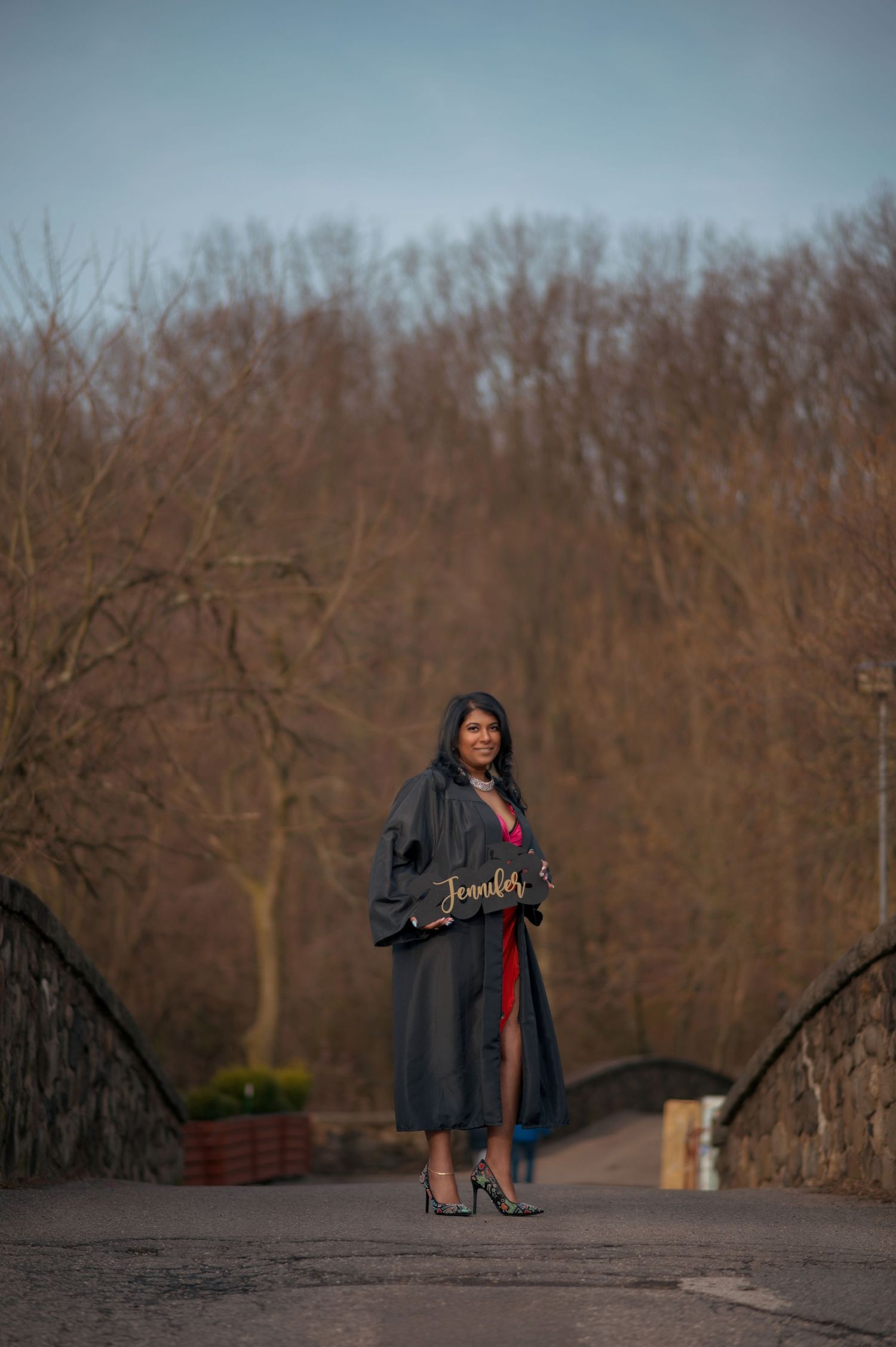 Graduate in black gown poses against winter landscape during sunset.