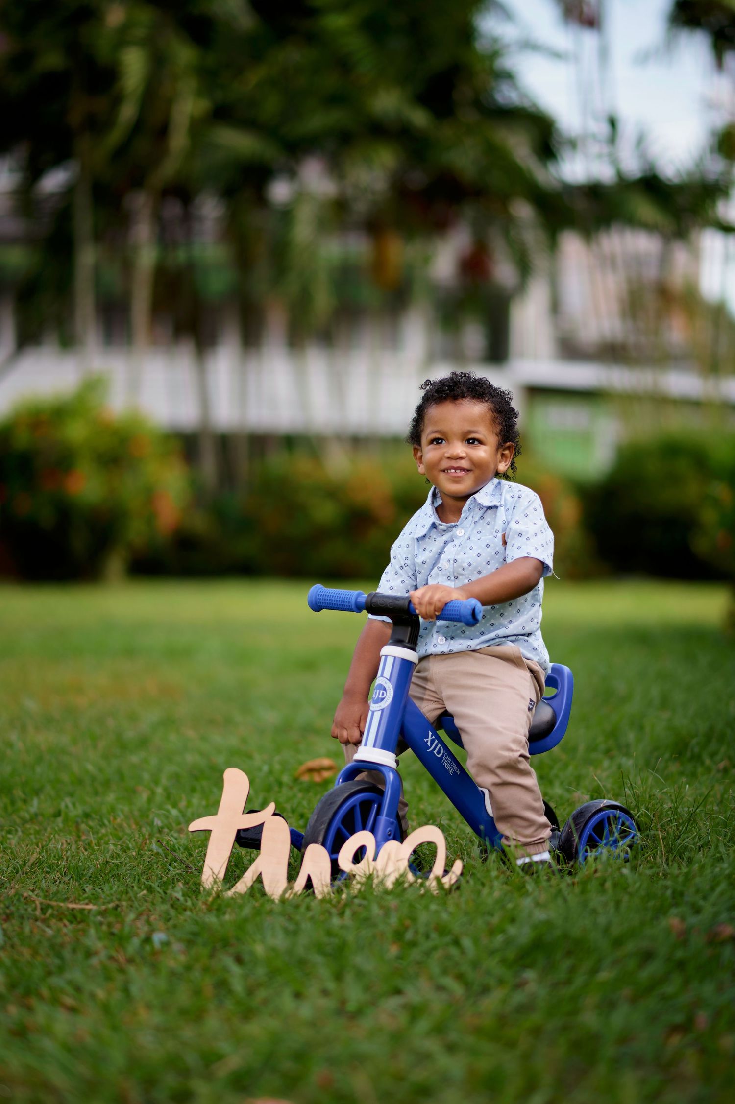 Small child sitting on a blue tricycle on grass with wooden decorative letters.