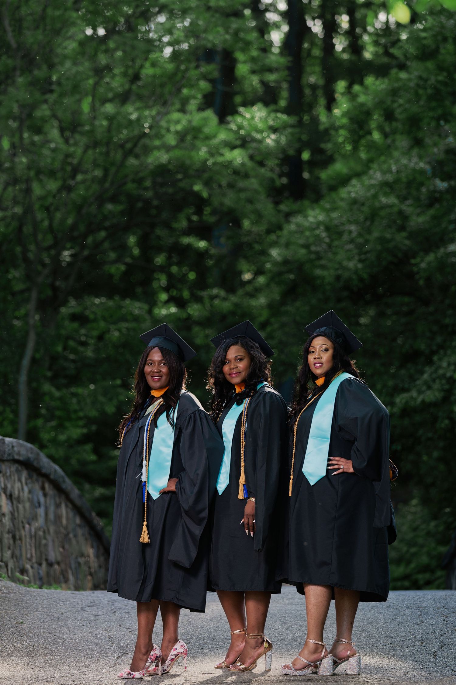 Three graduates in black gowns and light blue stoles pose together outdoors.