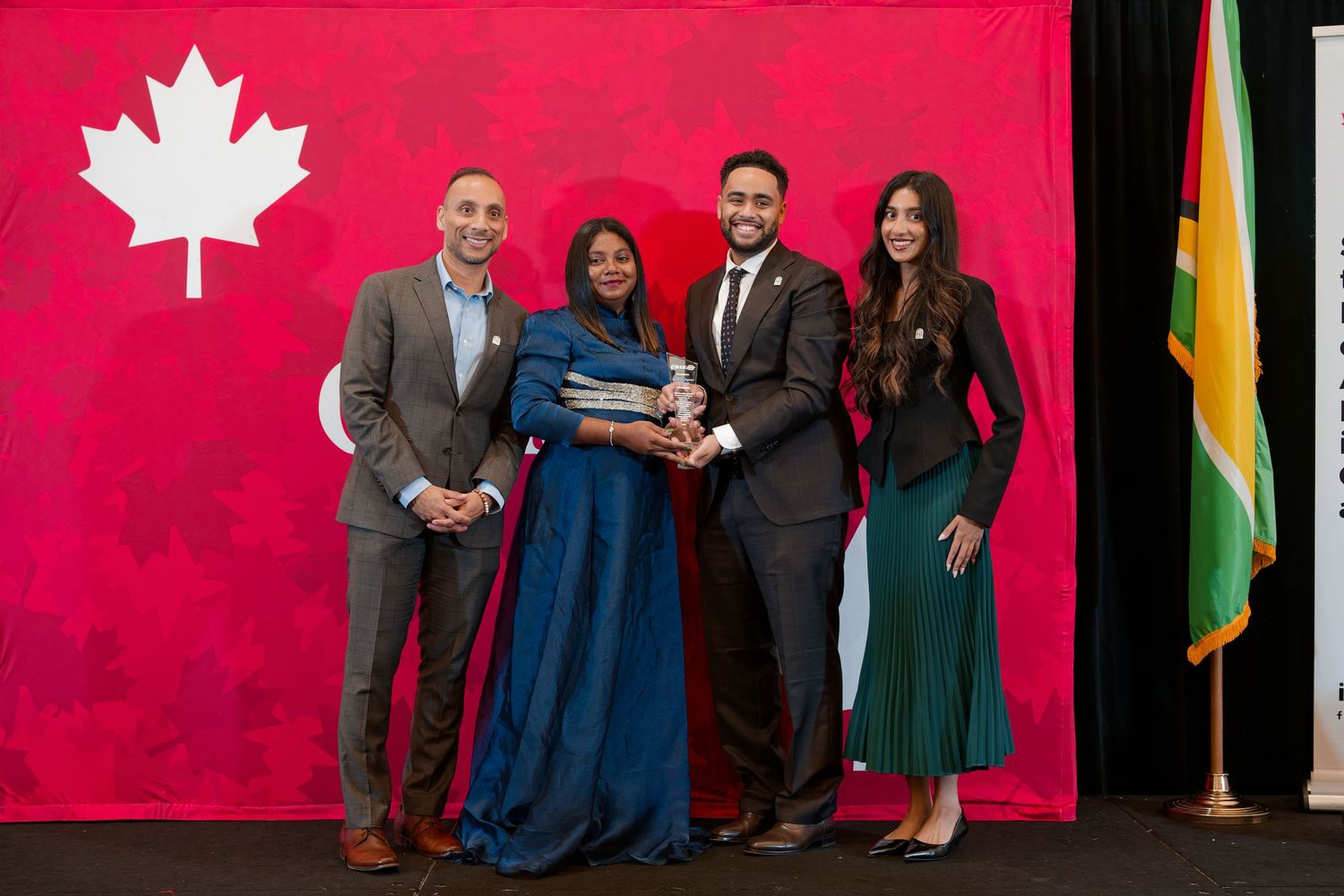 Four professionals pose with an award in front of Canadian and Guyanese flags at a formal ceremony.