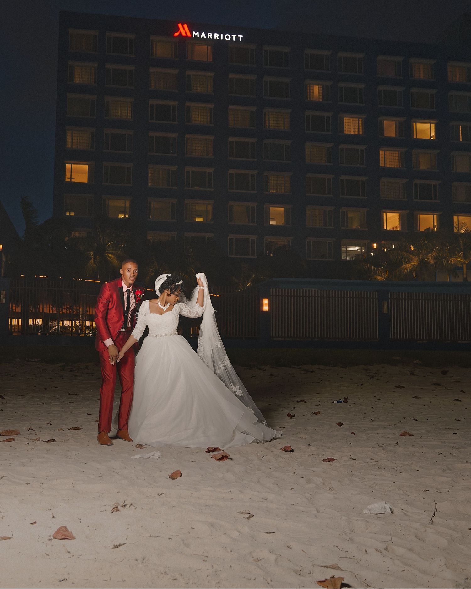 Bride and groom pose on sandy beach at night with illuminated Marriott hotel in background.