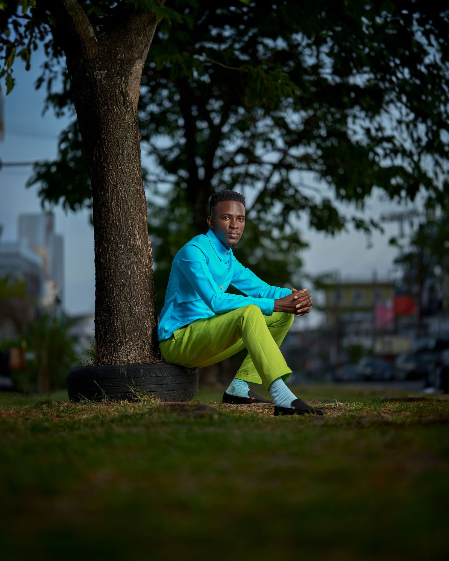 A man in a bright turquoise shirt and lime green pants sits under a tree in a park.