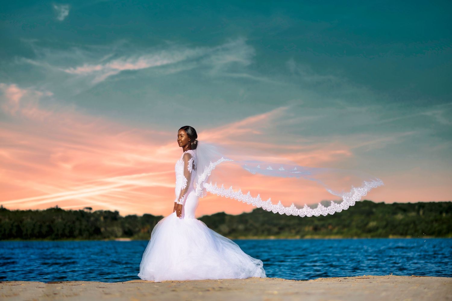 Bride in flowing white dress poses with veil flowing against waterfront sunset.