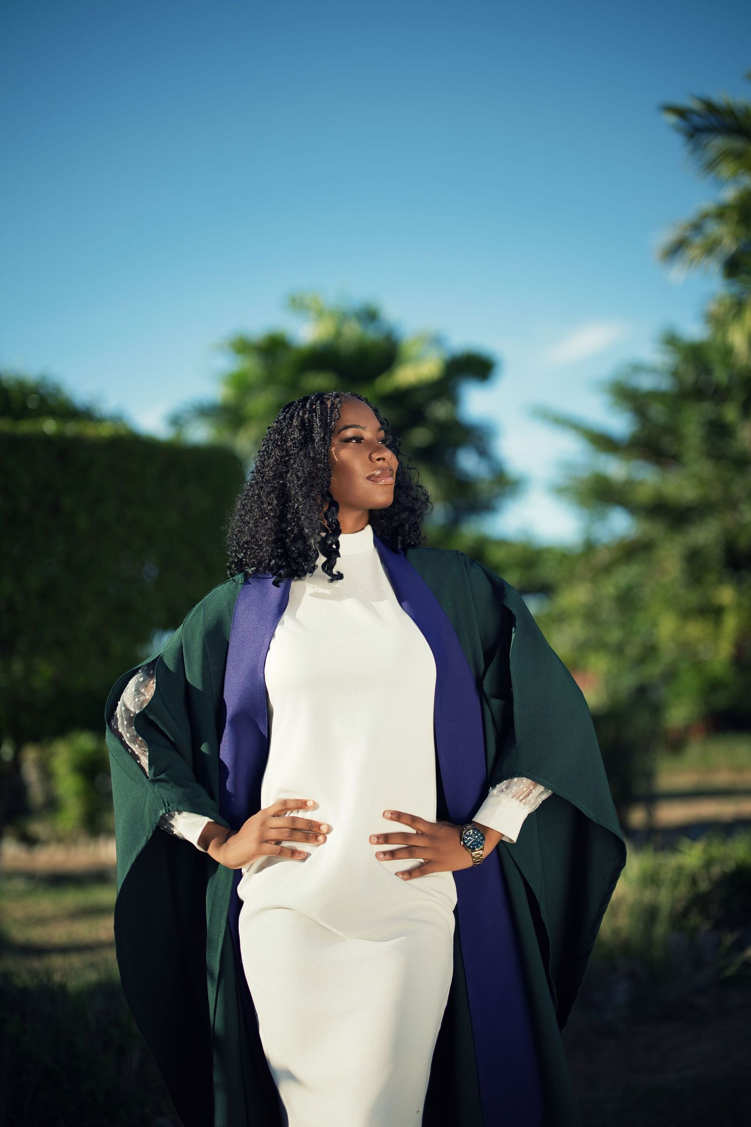 Graduate in white dress and dark robe poses against sunny outdoor backdrop.